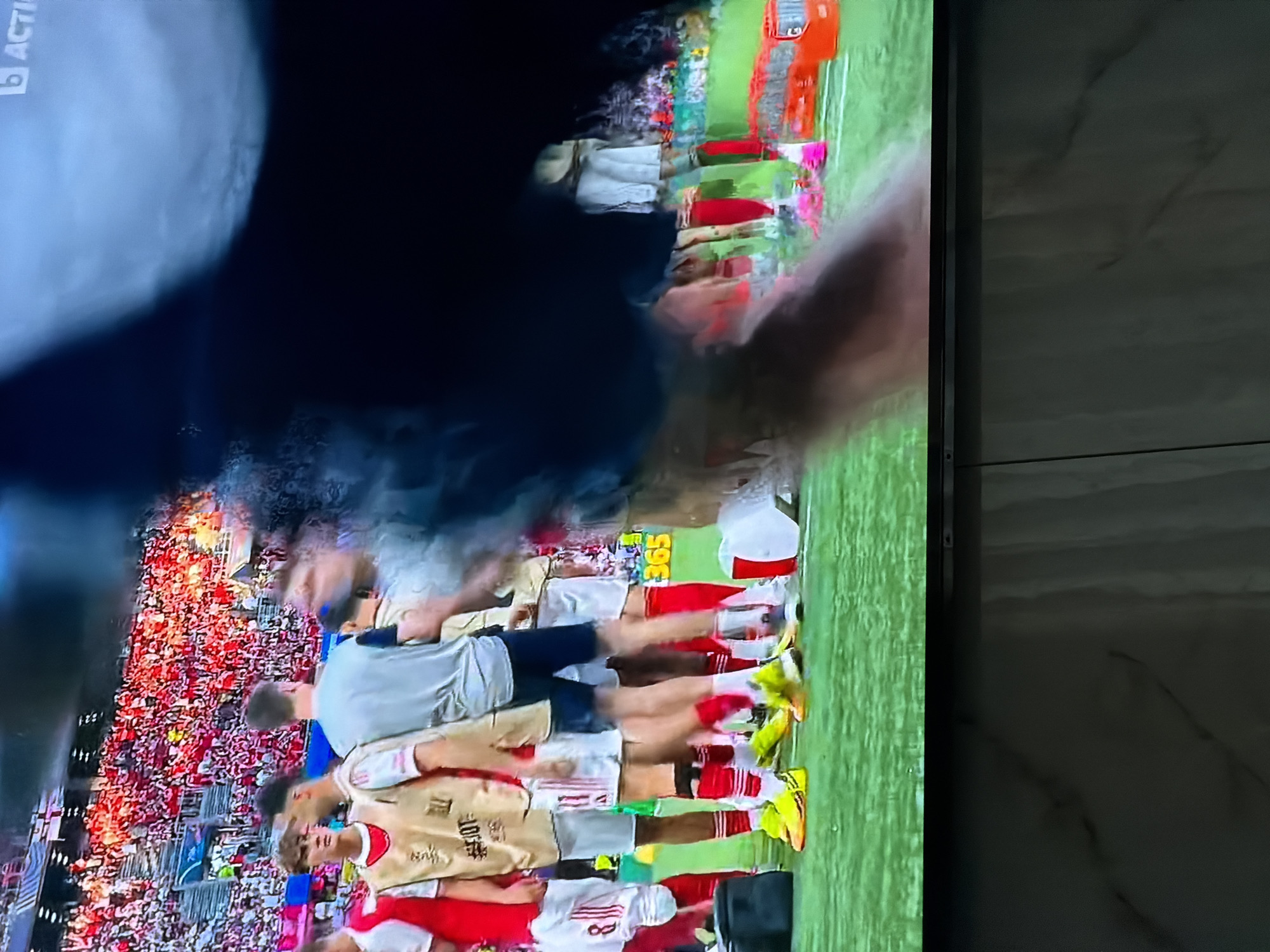 The image shows a television screen displaying a soccer game with a dense crowd in the stands. Players in red and white uniforms, some wearing gold training vests, are lined up on the green field. The background features advertisements and a green lawn. The exact location in Asenovgrad, Bulgaria, is not discernible from the image.