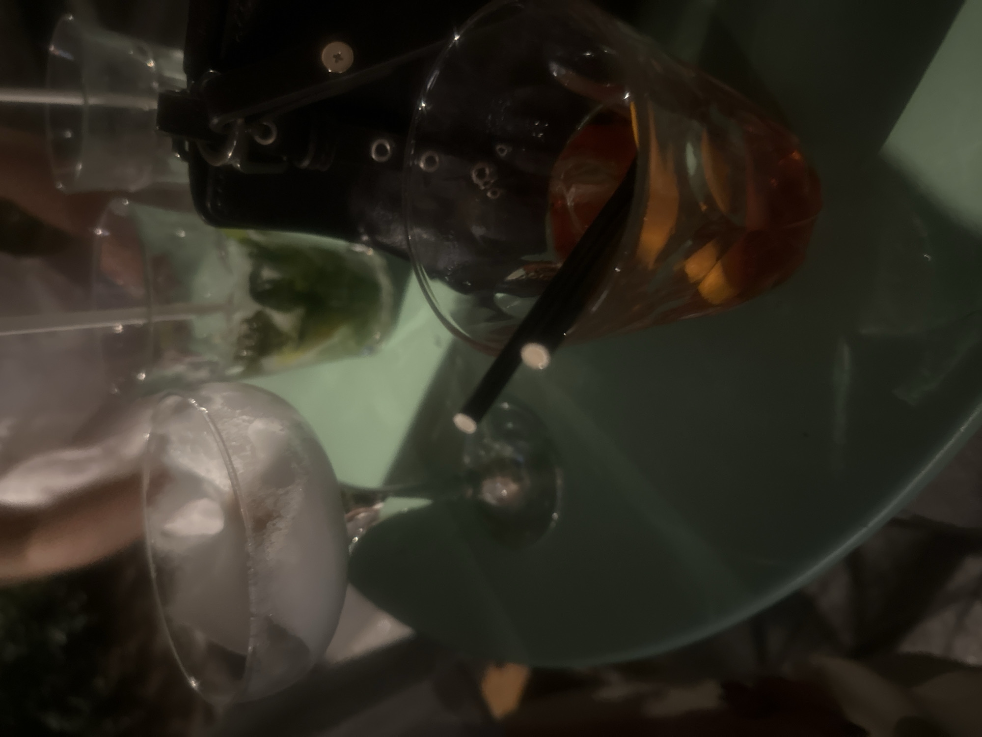 The image shows three drinks on a light green table. One drink contains ice, another appears to be a mojito, and the third is an orange-colored drink with a black straw. A black object with metal accents is also visible. The location and time of day are not discernible from the image.