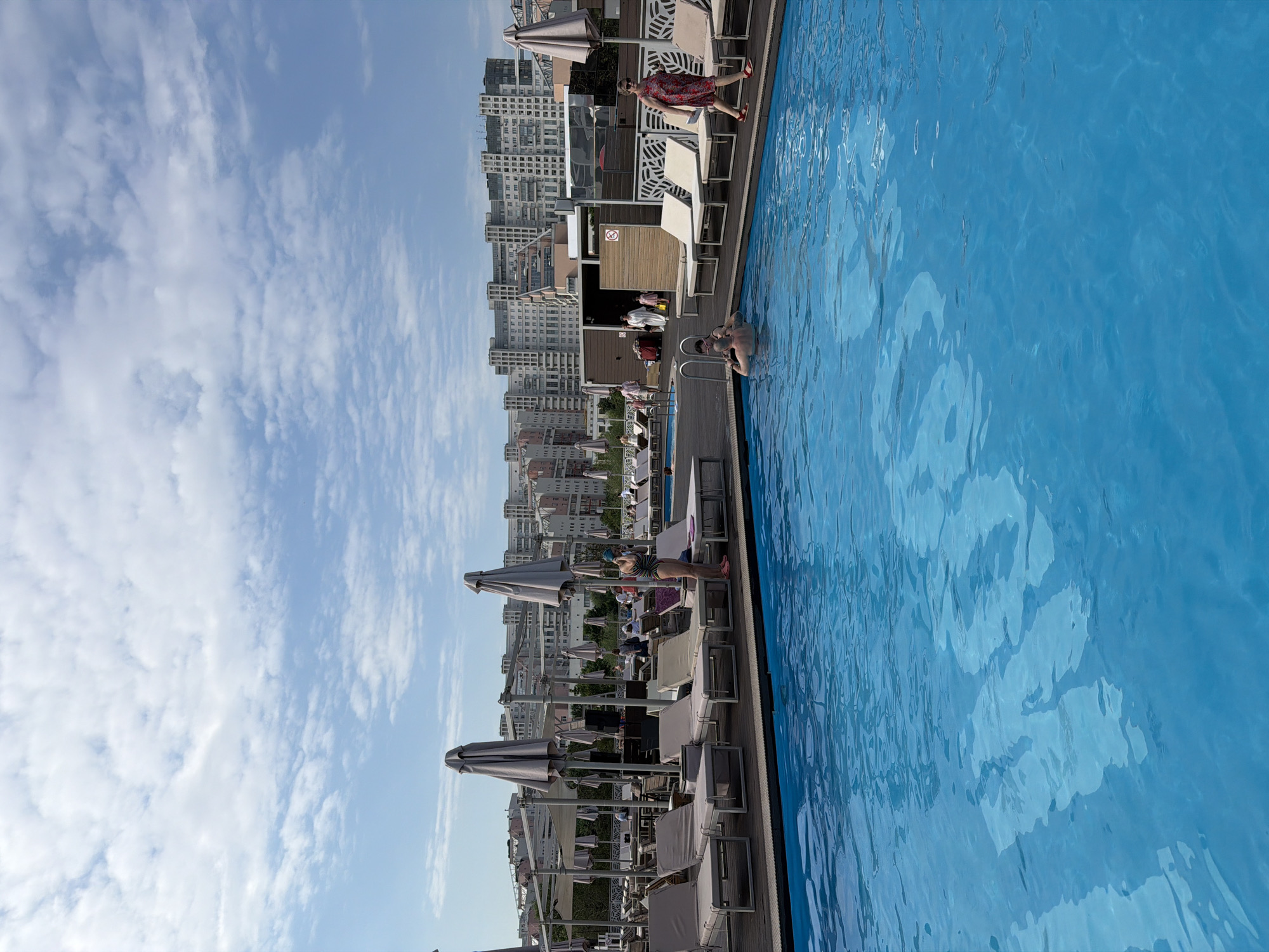 A rooftop swimming pool is depicted, situated in the city of Khimki, Russia, with a panorama of urban buildings in the background under a cloudy sky. The pool is filled with vibrant blue water, and the surrounding area features wooden decking, multiple lounge chairs, and large umbrella structures providing shade. Several individuals are visible. In the foreground, a man is partially submerged in the pool, his head and shoulders visible. Near the pool's edge, a woman in a striped swimsuit stands, holding a towel or similar item. Further away, another woman in a red floral dress walks along the poolside, and other patrons are seen relaxing on lounge chairs or walking in the background. The overall scene suggests a leisurely day at a poolside establishment.