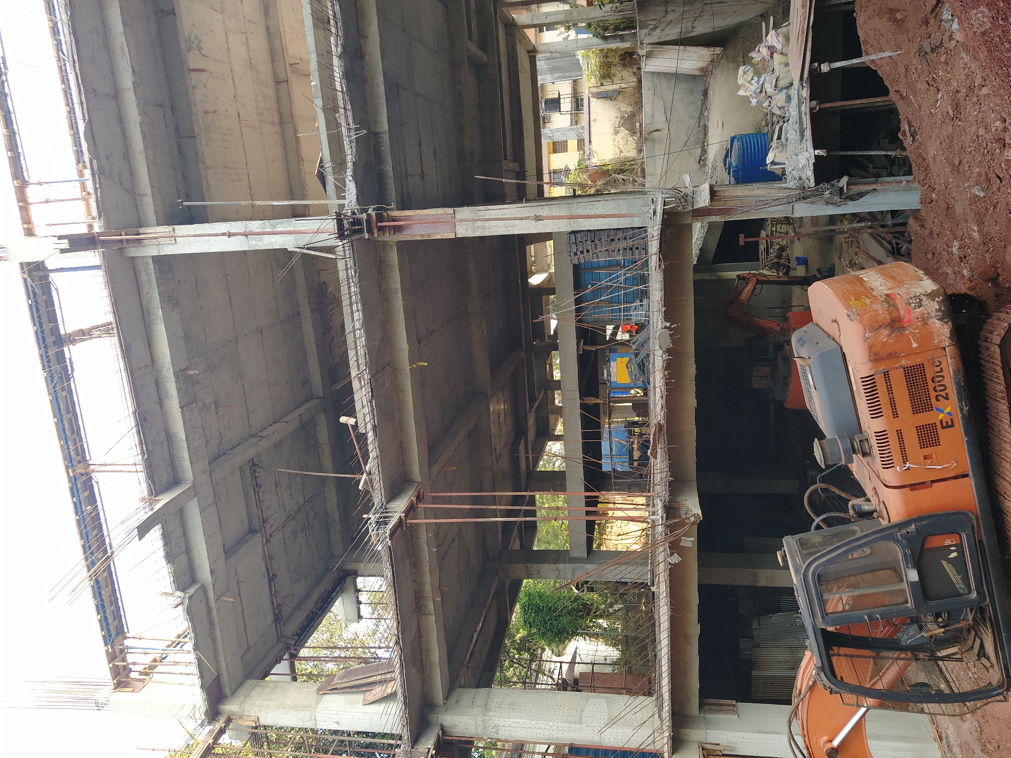 The image shows a construction site in Mapusa, India. A partially completed multi-story concrete building is visible, with exposed rebar and formwork. An orange excavator is parked on the right side of the frame, adjacent to a dirt pile and construction debris. In the background, partially obscured by the building and debris, are other buildings, suggesting an urban setting. The scene is typical of ongoing construction activities in a developing area.
