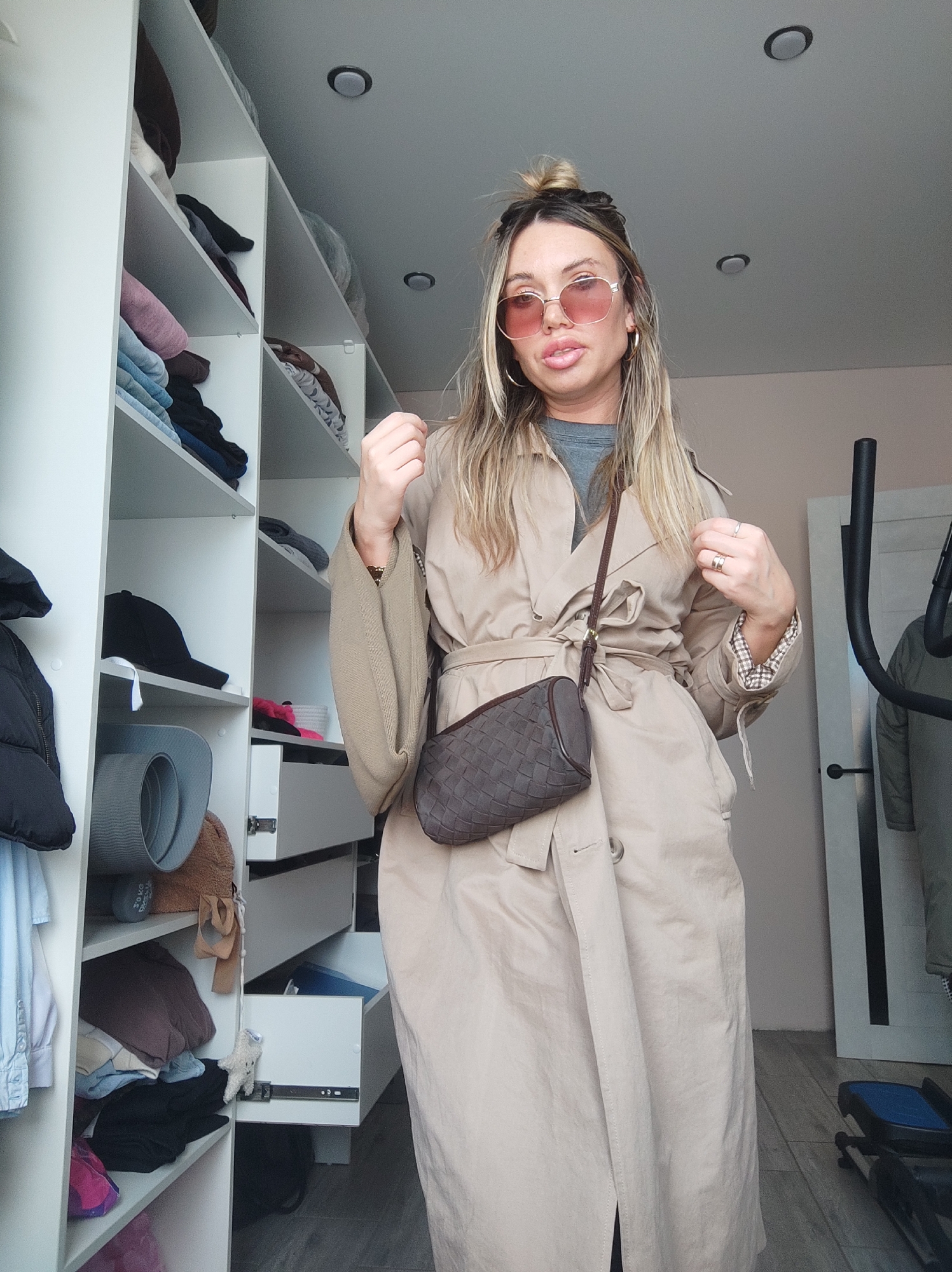 The image displays an adult female standing within what appears to be a domestic interior, likely a bedroom or dressing room. The woman, with blonde hair partially styled in a bun, wears pink-tinted, gold-rimmed sunglasses, a grey t-shirt, and a belted light beige trench coat with plaid lining visible at the cuffs. A dark brown woven crossbody bag is positioned across her body. She directly faces the camera, with her left hand raised towards her head and her right hand near her collar.
To the subject's left, a white modular storage unit or wardrobe system is visible, featuring multiple open shelves containing folded garments such as jeans, sweaters, and a black baseball cap. A grey rolled mat is also on a shelf. A black puffer jacket hangs on the extreme left. A lower drawer in the wardrobe is partially open, displaying a small white stuffed animal clipped to its edge.
On the right side of the frame, a light-colored wall is present, along with a white door featuring a dark vertical panel. A portion of a black metal object, consistent with exercise equipment, is visible behind the woman. The ceiling is light grey and contains two circular recessed light fixtures. The floor is covered with light-toned material, possibly wood or laminate, with a blue and black object resting on it near the door. The specific location within the city of Votkinsk, Russia, cannot be determined from the visual information provided.