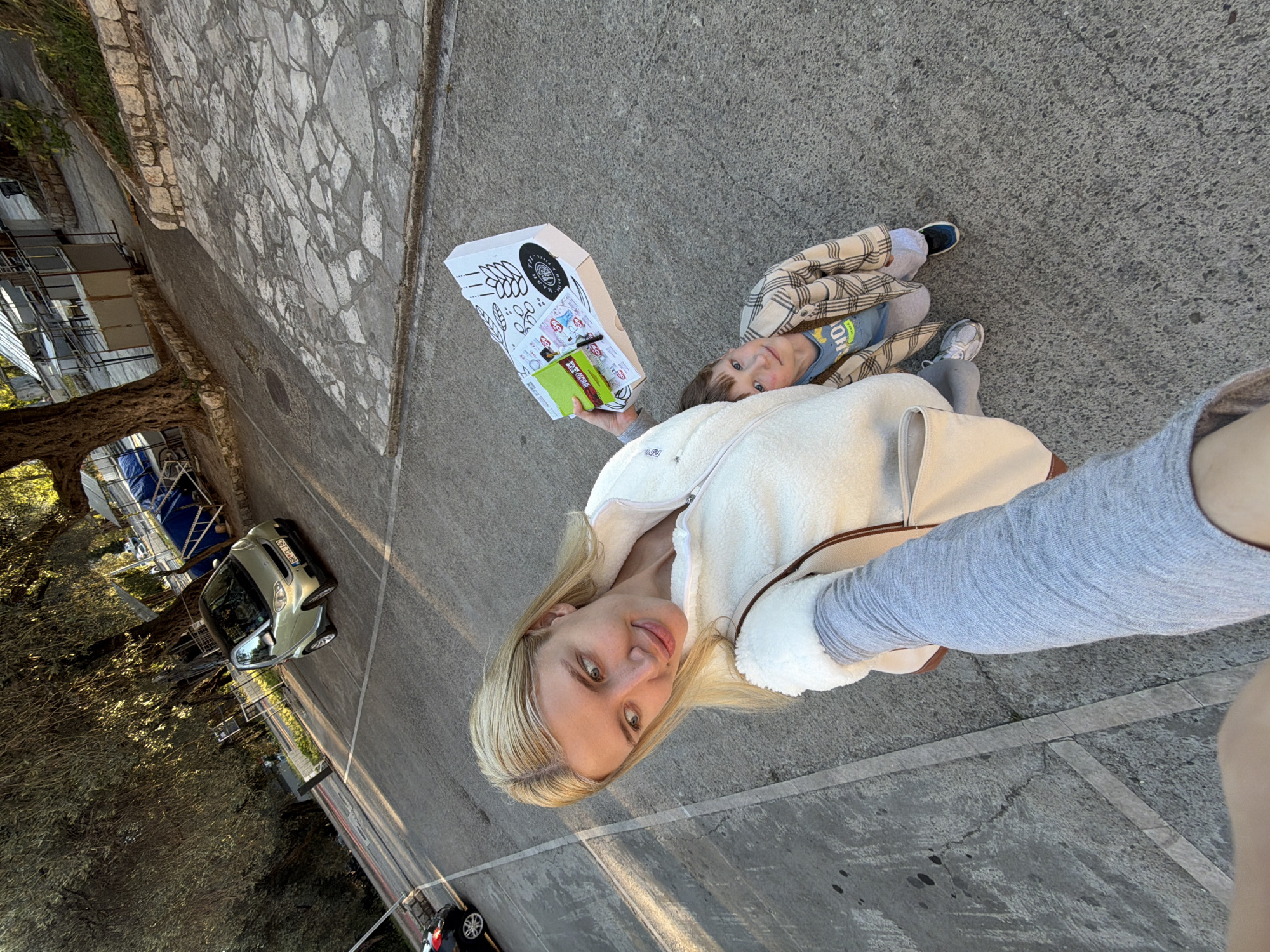 A light-skinned blonde woman with shoulder-length hair takes a selfie with a young child on a paved surface in Petrovac centar, Montenegro. The woman wears a white fleece jacket and a gray long-sleeved shirt. The child, wearing a plaid shirt, lays near her, looking up. She holds a white takeout box and a green package in one hand. The background includes a tree, parked cars, and buildings. The perspective is angled, showing parts of a street and sidewalk.
