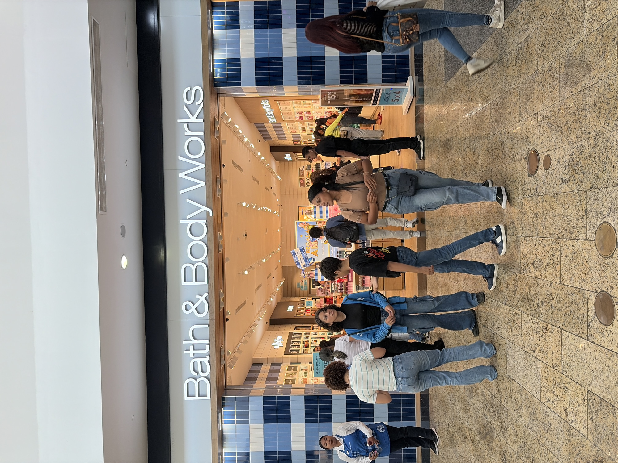 The image shows the exterior of a Bath & Body Works store, with several people standing in front of it. The store has a white facade with blue and white checkered tiles at the bottom. The Bath & Body Works logo is prominently displayed above the entrance. People of varying ages are standing on a tan-colored, tiled floor in front of the store. The interior of the store is visible, revealing shelves stocked with products. The scene is likely inside a shopping mall. There is no indication of being located in the city of Ciudad Guayana, Venezuela.
