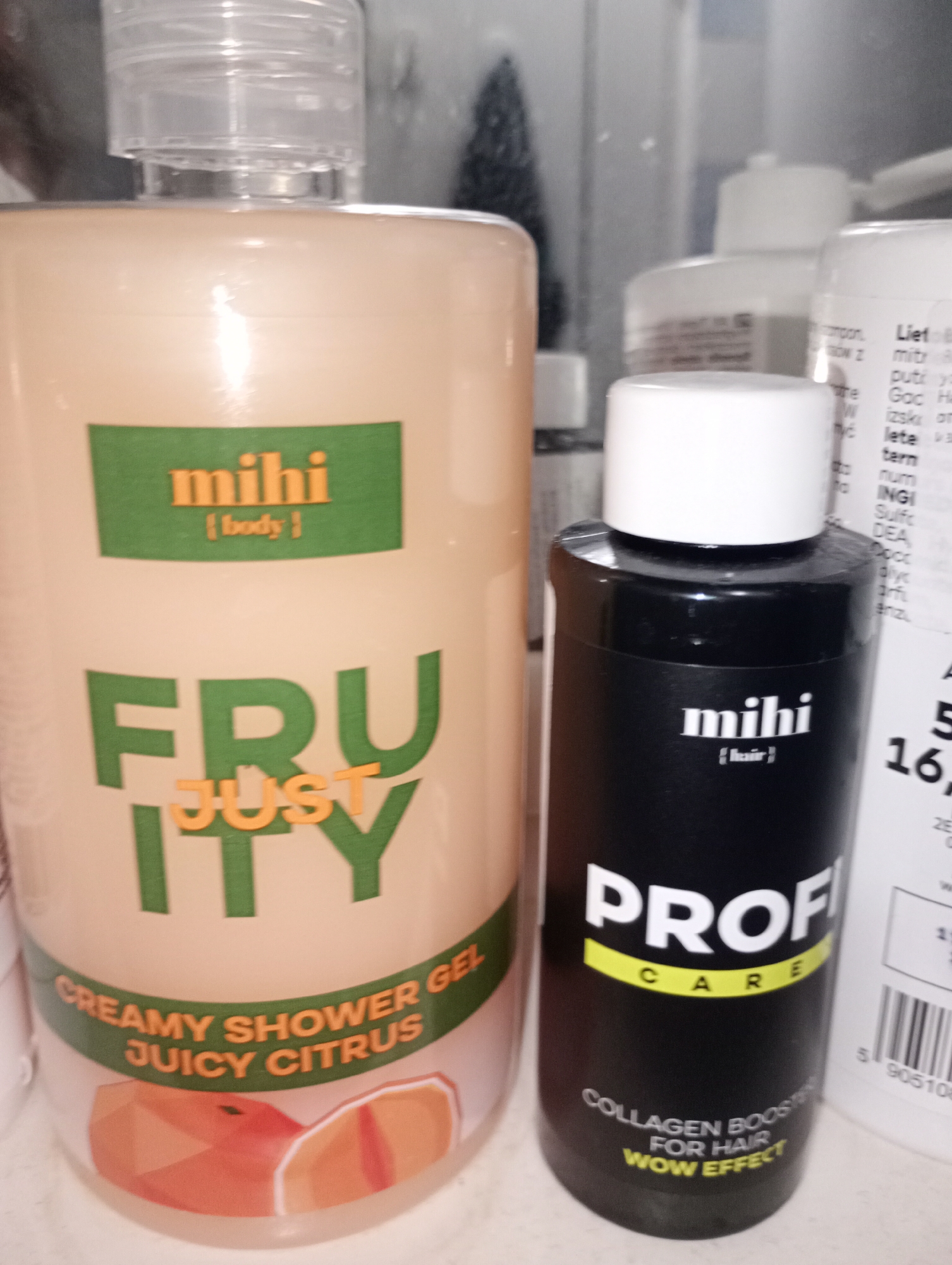 In the city of Harmanli, Bulgaria, an indoor photograph captures a collection of cosmetic products, primarily shampoos and shower gels, arranged on a shelf. The foreground is dominated by two large bottles. On the left, a tall, pale orange bottle with a clear plastic cap displays the "mihi {body}" branding in green and orange. Below this, the words "FRUITY JUST" are prominently featured in large green letters, with "JUST" in orange. The lower section of this bottle is adorned with a curved label that reads "CREAMY SHOWER GEL JUICY CITRUS" and features an illustration of sliced citrus fruits. To the right of the shower gel, a smaller, matte black bottle with a white cap is visible. This bottle also carries the "mihi {hair}" branding in white. Below this, the text "PROFI CARE" appears in white and yellow, followed by "COLLAGEN BOOST FOR HAIR WOW EFFECT" in white lettering. Partially visible in the background are other cosmetic containers, including white bottles with text and what appears to be a barcode sticker. The lighting suggests an indoor setting, likely a retail or home environment, with the products neatly arranged.