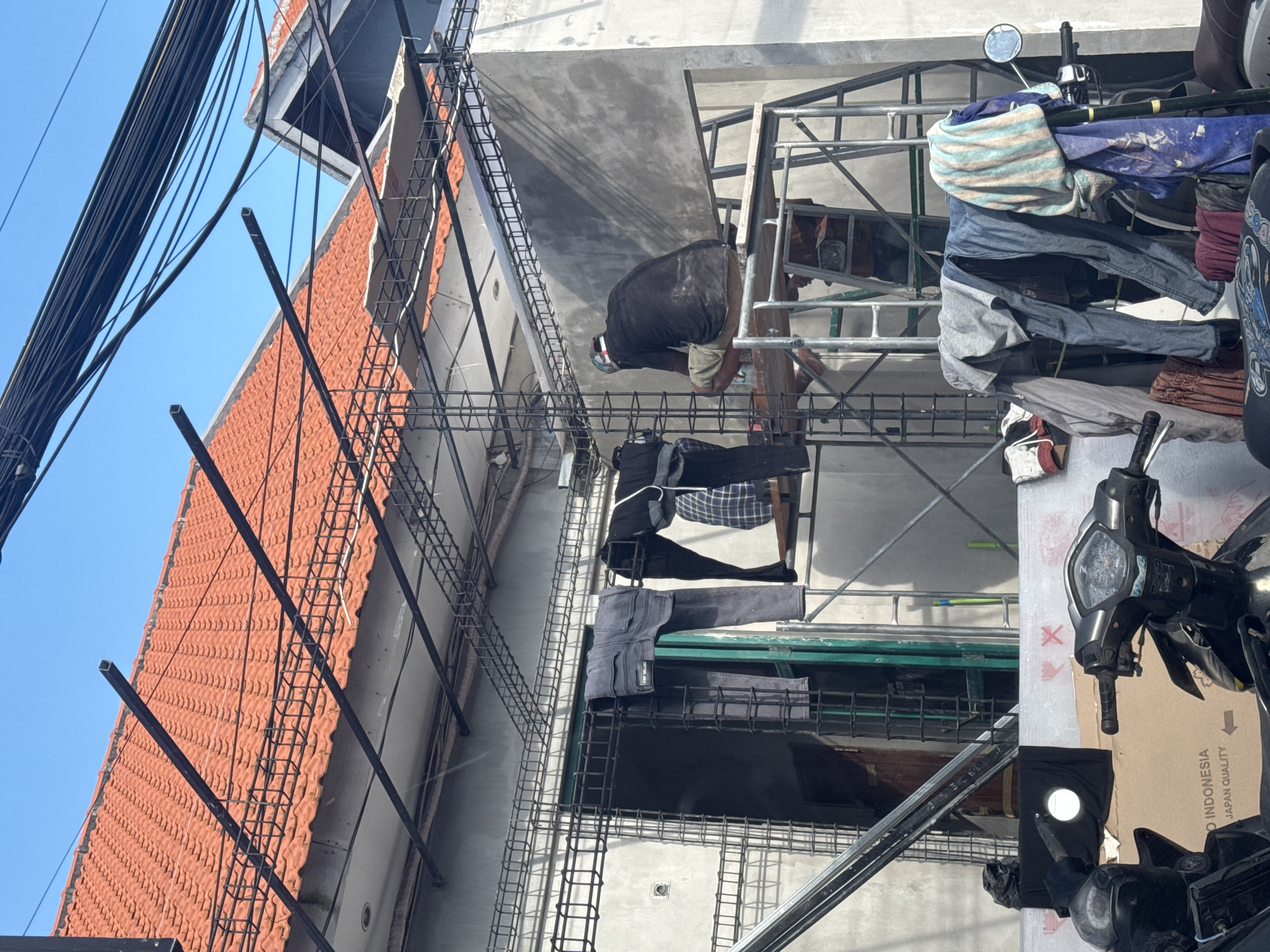 The image depicts a construction site in North Kuta, Indonesia. The facade of a building under construction is visible, featuring an orange tile roof, concrete walls, and metal rebar reinforcement. Scaffolding surrounds the building, with a construction worker visible on the upper level. Clothes are hanging to dry on the scaffolding. A parked motorcycle is on the right side of the frame. Power lines run across the top left corner. The sky is visible in the background.