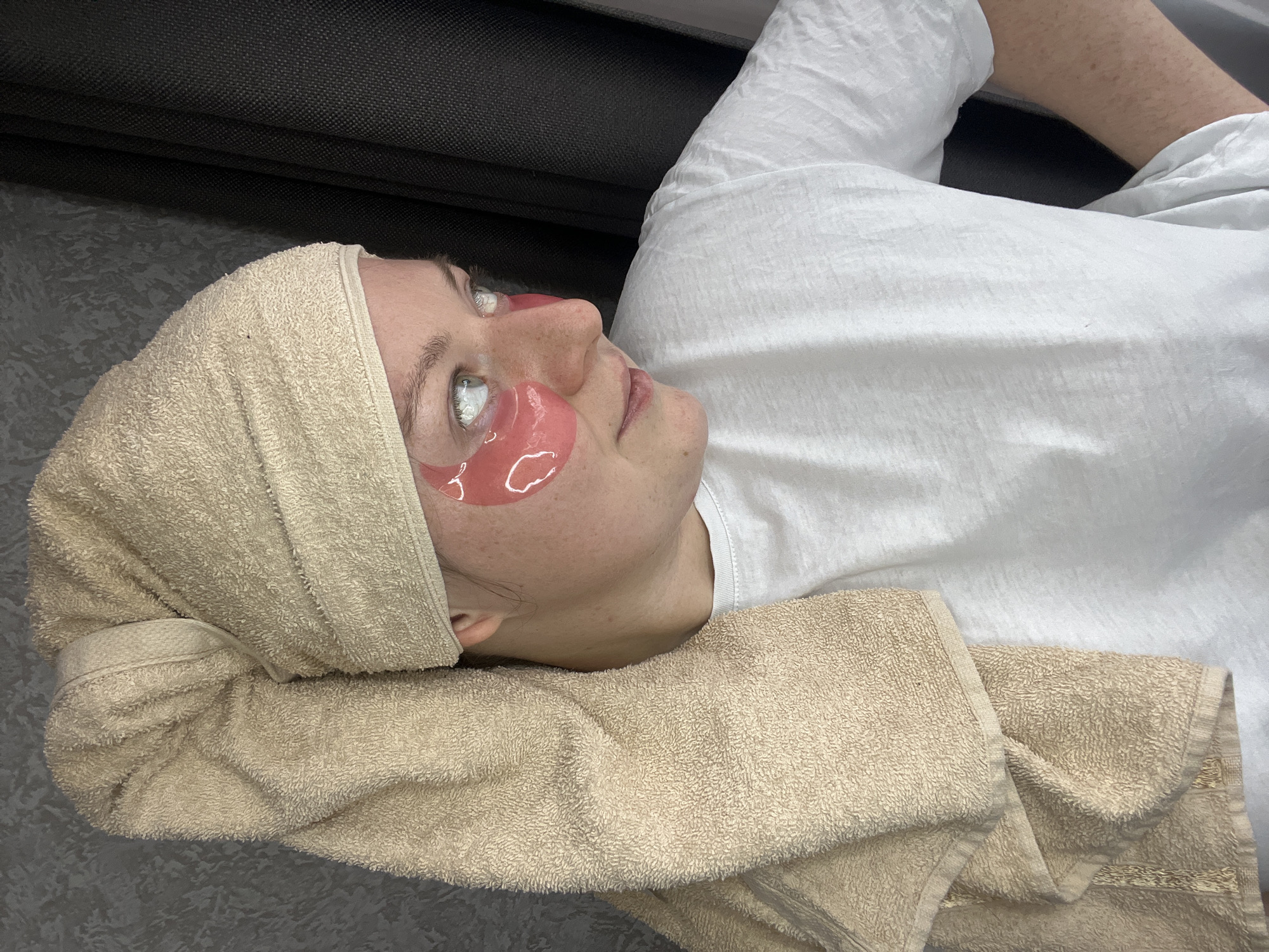 A woman with light skin is lying on her back wearing pink under-eye patches and a light-beige towel wrapped around her head. A folded beige towel supports her head. She's wearing a white t-shirt, and part of a gray sofa is visible in the upper-left. The floor is gray. The image was taken indoors. There's no information indicating the location is Раздолье, Russia.