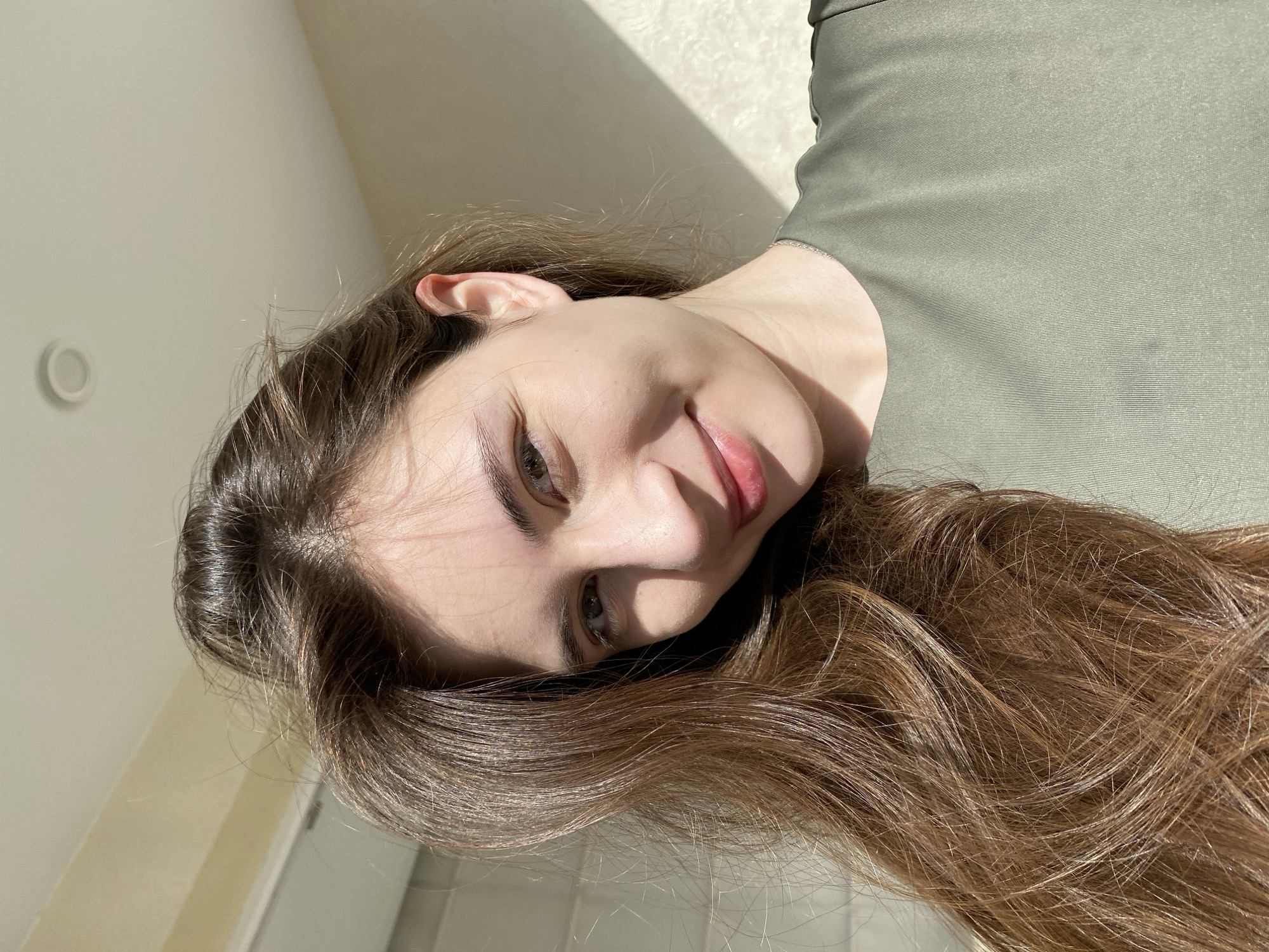 A close-up, angled shot of a fair-skinned woman with long, light brown hair, hazel eyes, and a slight smile. She is wearing an olive green top. The background includes a light-colored wall partially illuminated by sunlight. There is no specific evidence in the image to confirm a location in Votkinsk, Russia.