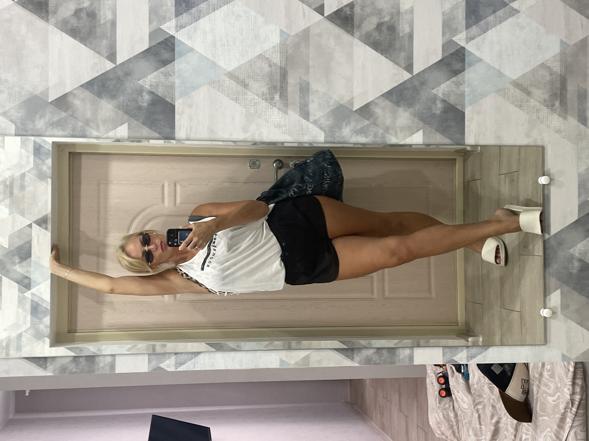A woman with blonde hair and sunglasses is taking a selfie in a hallway, leaning against a door frame. She is wearing a white tank top and black shorts. A denim bag is slung over her shoulder. The walls are decorated with a geometric pattern of grey, white, and blue triangles. In the reflection of the mirror, a bed with a patterned duvet and a baseball cap can be seen. The scene appears to be an indoor residential setting, likely an apartment in Novorossiysk, Russia, given the context of the request.