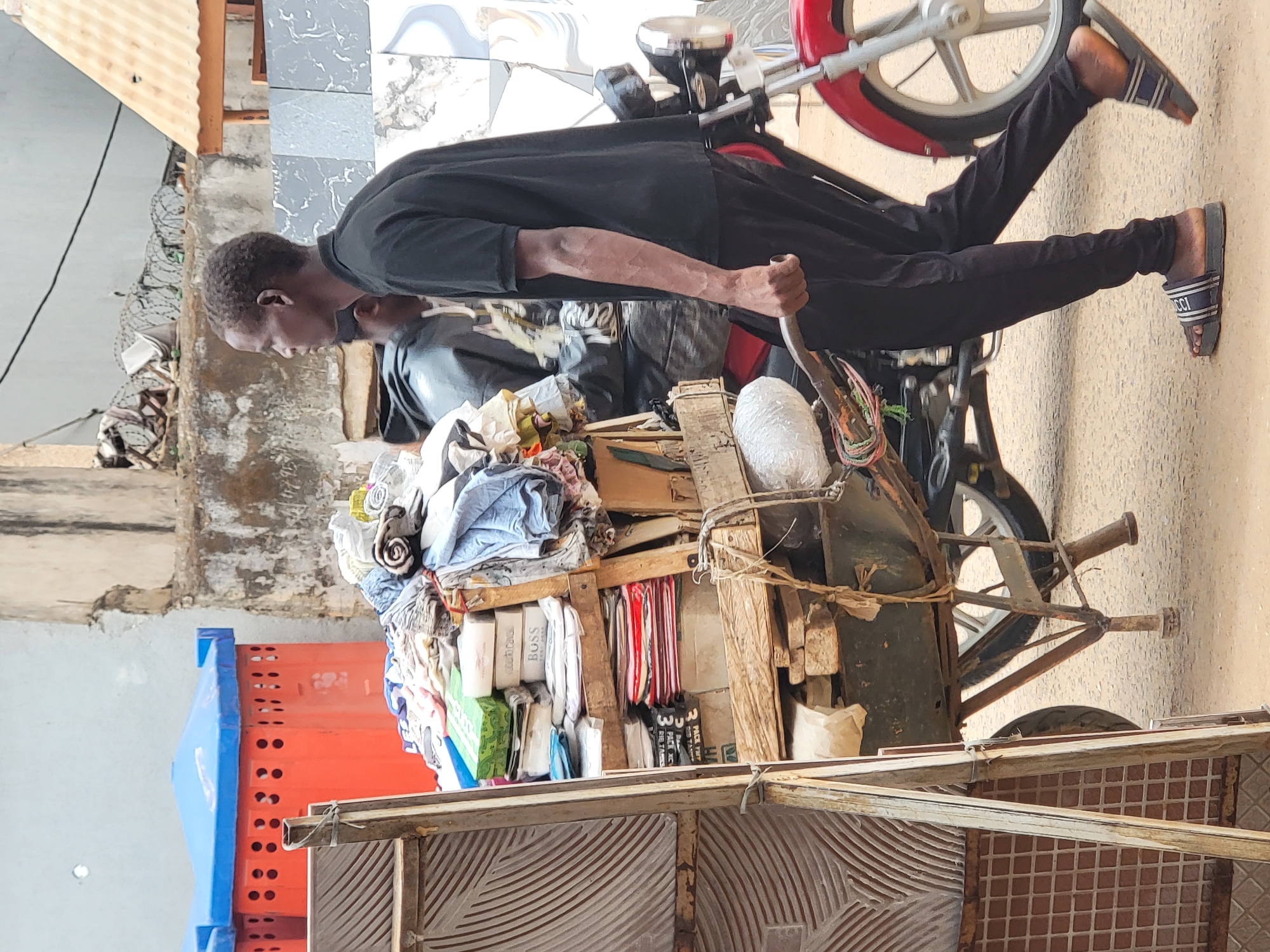 A man in Maiburiji, Nigeria, pulls a heavily laden cart. The cart is constructed of wood and metal, and is stacked high with textiles and other goods. The man is wearing dark clothing and sandals. The environment appears to be a street with buildings in the background. The scene is captured during the day.