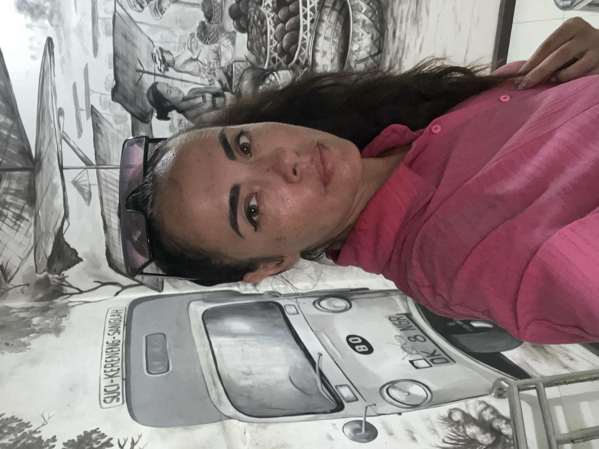 A woman with long, dark hair and sunglasses on her head takes a selfie in front of a mural. The mural depicts a street scene with a three-wheeled vehicle and market stalls. The woman wears a pink shirt. The mural is painted in black and white, with some muted colors in the baskets. The text on the mural reads "SUCI-KERENENG-SANGLAH" and "DK 8 NGR". The location is identified as Kuta Selatan, Indonesia.