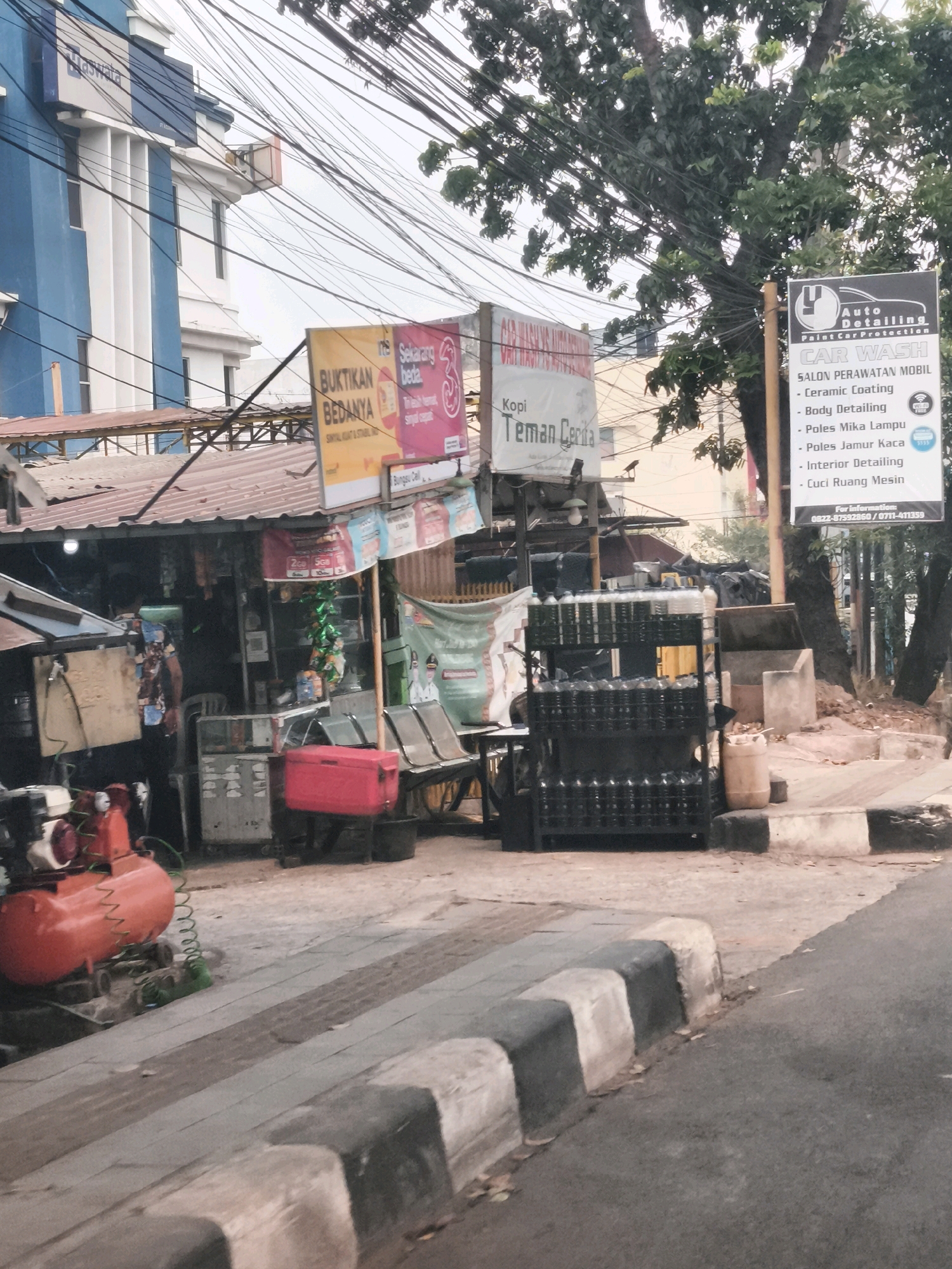 The image shows a street scene in Palembang, Indonesia, with a small shop selling various items and a car wash service. On the left side, a blue and white building with a sign that says "Haswara" is partially visible. In front of the shop, an orange air compressor is placed on the sidewalk. The shop itself has a corrugated metal roof and displays various banners and signs advertising services and products. One banner prominently advertises "Kopi Teman Cerita" (Coffee Friend Story), indicating a cafe. Another sign promotes "Auto Detailing" and "Car Wash" services, listing specific offerings such as "Ceramic Coating", "Body Detailing", "Poles Mika Lampu", "Poles Jamur Kaca", "Interior Detailing", and "Cuci Ruang Mesin". This car wash sign also includes contact information. Several rows of plastic bottles are stacked on a black metal rack in front of the shop. In the background, trees and utility wires are visible, typical of an urban environment. The street has a concrete sidewalk with a curb marked with alternating black and white sections. The overall scene depicts a busy commercial area with a mix of small businesses.