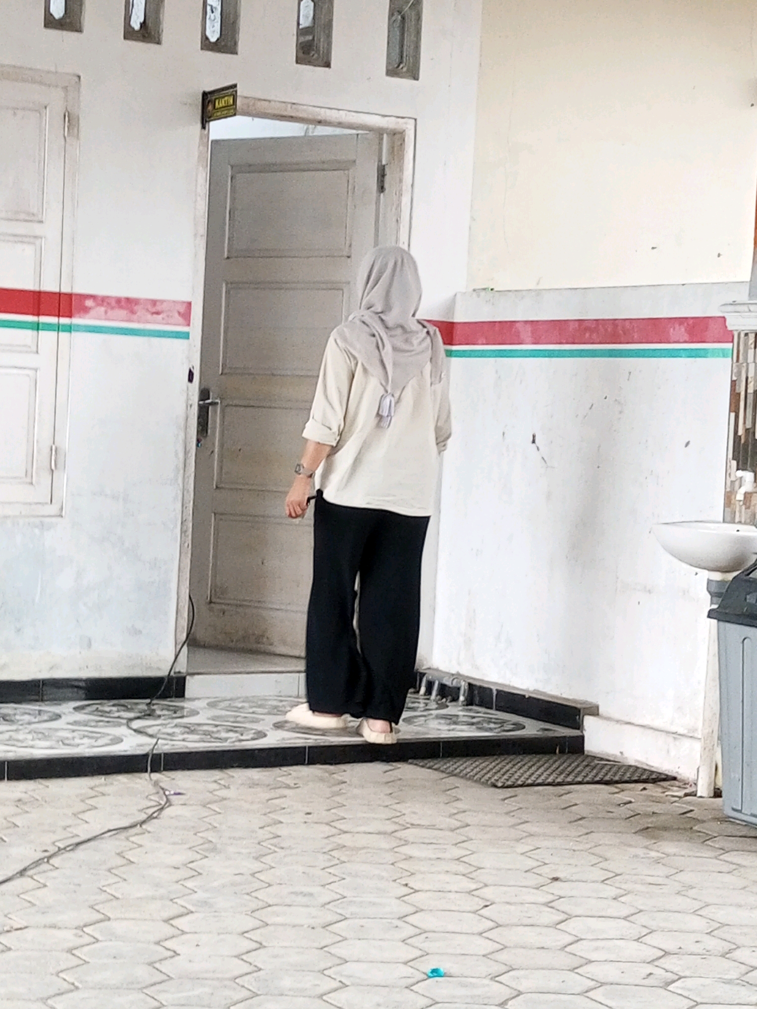 A woman wearing a light-colored hijab and a cream-colored blouse over black pants stands in front of an open door with a sign that reads "KANTIN" in Indonesian. The woman is facing away from the camera, and appears to be walking or about to enter the room. The building has white walls with red and green horizontal stripes. The ground in front of the building is paved with hexagonal tiles, and there is a black doormat to the right of the doorway. A white sink is visible on the right side of the image. The scene is located in Kalibeber, Indonesia.