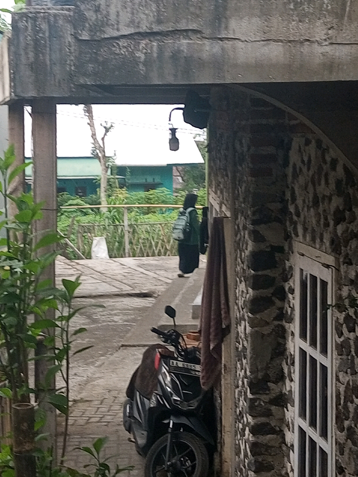 In Andongsili, Indonesia, a person wearing a dark garment and a light-colored backpack walks away from the camera on a concrete walkway. The walkway is bordered by a low wooden fence and lush green foliage. Behind the fence, a teal building with a white roof is visible, suggesting a residential or institutional setting. A motorcycle is parked on the right side of the walkway, partially covered with a brown cloth. The scene is framed by a rough concrete structure in the foreground and a textured stone wall with a white-framed window on the right. A light fixture hangs from the concrete structure, and a thin branch from a tree is visible in the background.