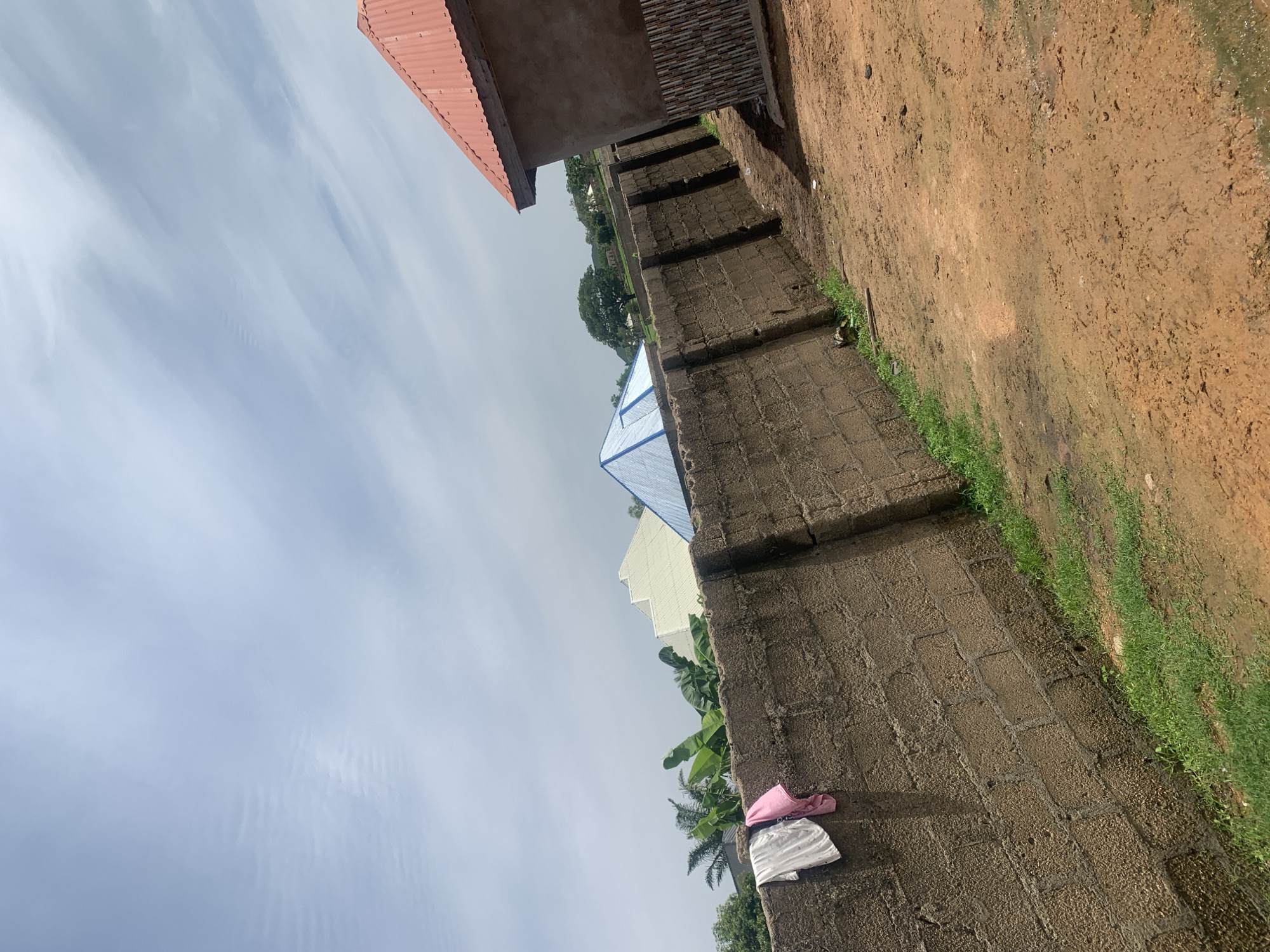 The image shows an outdoor scene, likely in a rural or semi-urban area, with a focus on a concrete block wall. Two pieces of clothing, a white shirt and a pink garment, are hanging on the wall. Behind the wall, several structures are visible, including buildings with corrugated metal roofs, one in red and another with a blue pyramidal roof. There are also trees and vegetation, including banana leaves, in the background. The ground in the foreground is a reddish-brown dirt, appearing somewhat uneven and wet in places. The sky is overcast with scattered clouds. The scene is located in Jauro Yinu, Nigeria.