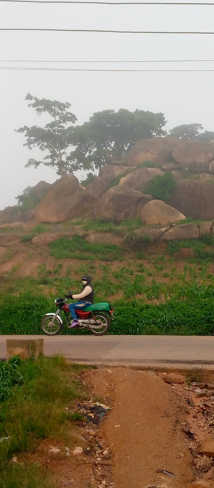 This media file depicts a person riding a motorcycle on a paved road. The rider is wearing a dark vest over a light-colored long-sleeved shirt, blue jeans, and purple shoes. They are also wearing a face mask and a hood, suggesting it might be a cool or dusty day. The motorcycle is red and has a green bag secured to the back.
The scene is set against a backdrop of a rocky hill covered in sparse vegetation and trees. The sky is overcast and hazy, indicating potentially foggy or misty weather. In the foreground, the road is bordered by grassy areas and some scattered debris. The overall impression is of a rural or semi-rural environment.
The time of day is difficult to determine precisely due to the overcast sky, but the lighting suggests it is daytime. There is no visible text in the image. The mood is neutral, focusing on the act of travel and the natural landscape. The composition highlights the solitary rider against the vastness of the natural elements.