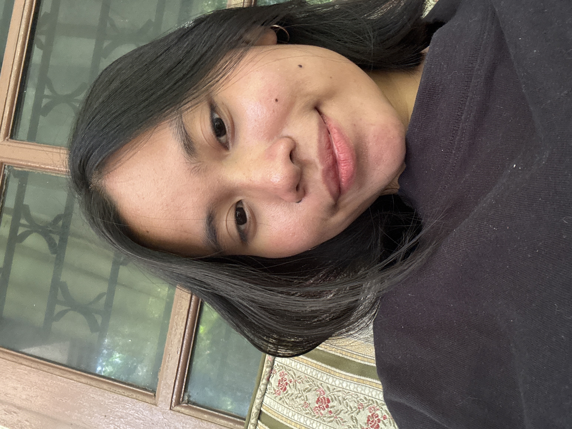 The image is a close-up selfie of a young woman with long, dark hair, smiling slightly. She is wearing a black top. Behind her, a window with wooden frames and decorative metalwork is visible, and a patterned sofa can be seen to the left. The scene appears to be indoors, possibly in a home or cafe, with natural light filtering through the window, suggesting it is daytime. The image does not contain any visual information that can definitively place it in South Jakarta, Indonesia.