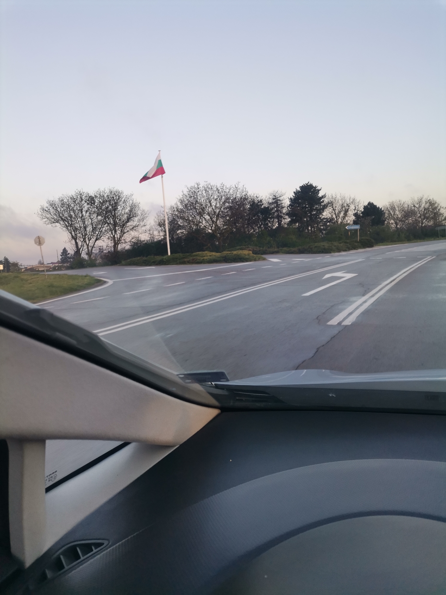 The image, captured from inside a vehicle, presents a view of a road junction or turn-off in Stefanovo, Bulgaria. A paved road stretches into the distance, featuring double solid white lines in the center and a prominent right-turn arrow painted on the right lane. On the left side of the road, a tall white flagpole displays the Bulgarian flag, characterized by its horizontal white, green, and red stripes, acting as a clear national identifier.
Bare-branched deciduous trees, indicative of late autumn, winter, or early spring, line both sides of the road and extend into the background, interspersed with some evergreen foliage. The sky above is light and overcast, suggesting either early morning, late afternoon, or simply a cloudy day, contributing to the soft, diffused light across the scene. The road surface appears damp, possibly from recent rain.
To the right of the road, partially obscured by trees, a blue directional sign with white text is visible, pointing towards "Шумен" (Shumen) and "Община" (Municipality), indicating the direction to the city of Shumen. In the foreground, parts of the car's interior, including the dashboard and windshield pillars, frame the outdoor view, confirming the perspective from within a vehicle. No specific activity beyond driving is depicted.