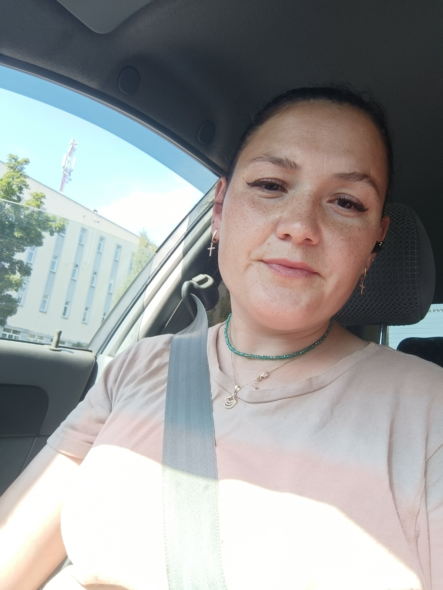 A woman is seated in the driver's seat of a car, looking at the camera. She has light brown hair pulled back, freckles across her face, and is wearing a light-colored t-shirt, a beaded necklace, and earrings. A seatbelt is visible across her chest. The background shows a building and trees through the car window. The image appears to be taken in the city of Chaikovsky, Russia.