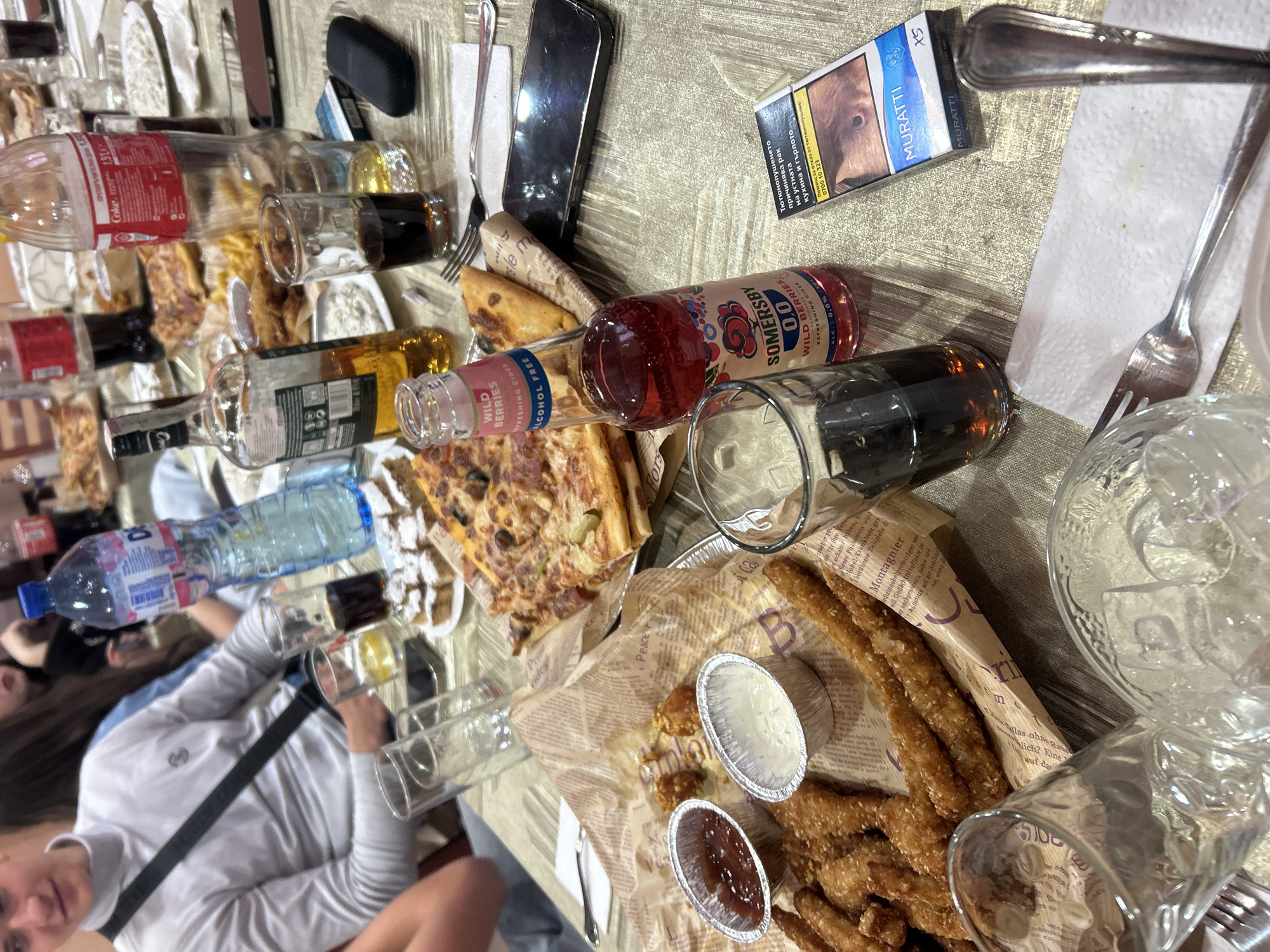 The image shows a table covered in various food and drink items, along with personal belongings, at what appears to be a social gathering.
**People:** Parts of at least two people are visible in the lower left corner. One person is wearing a white long-sleeved shirt.
**Subjects:** The primary subjects are food and drink items on the table.
**Objects:**
* Bottles: Coca-Cola, water, Somersby cider, and an unidentified brown liquid in a clear bottle.
* Food: Pizza, breaded sticks with dipping sauces.
* Glasses: Several glasses, some containing dark liquids.
* Personal items: A smartphone, a sunglasses case, and a pack of Muratti XS cigarettes.
* Cutlery: Forks and knives.
* Paper napkins.
**Actions/Interactions:** Indeterminate
**Environment:** The scene is indoors, on a table. The lighting appears to be artificial. The objects are cluttered and suggest a recent or ongoing meal.
**Location:** The presence of "0700 10 323" on the cigarette pack could suggest that the location is within Bulgaria. However, there is no direct visual confirmation of the location in Mirkovo, Bulgaria in this image.