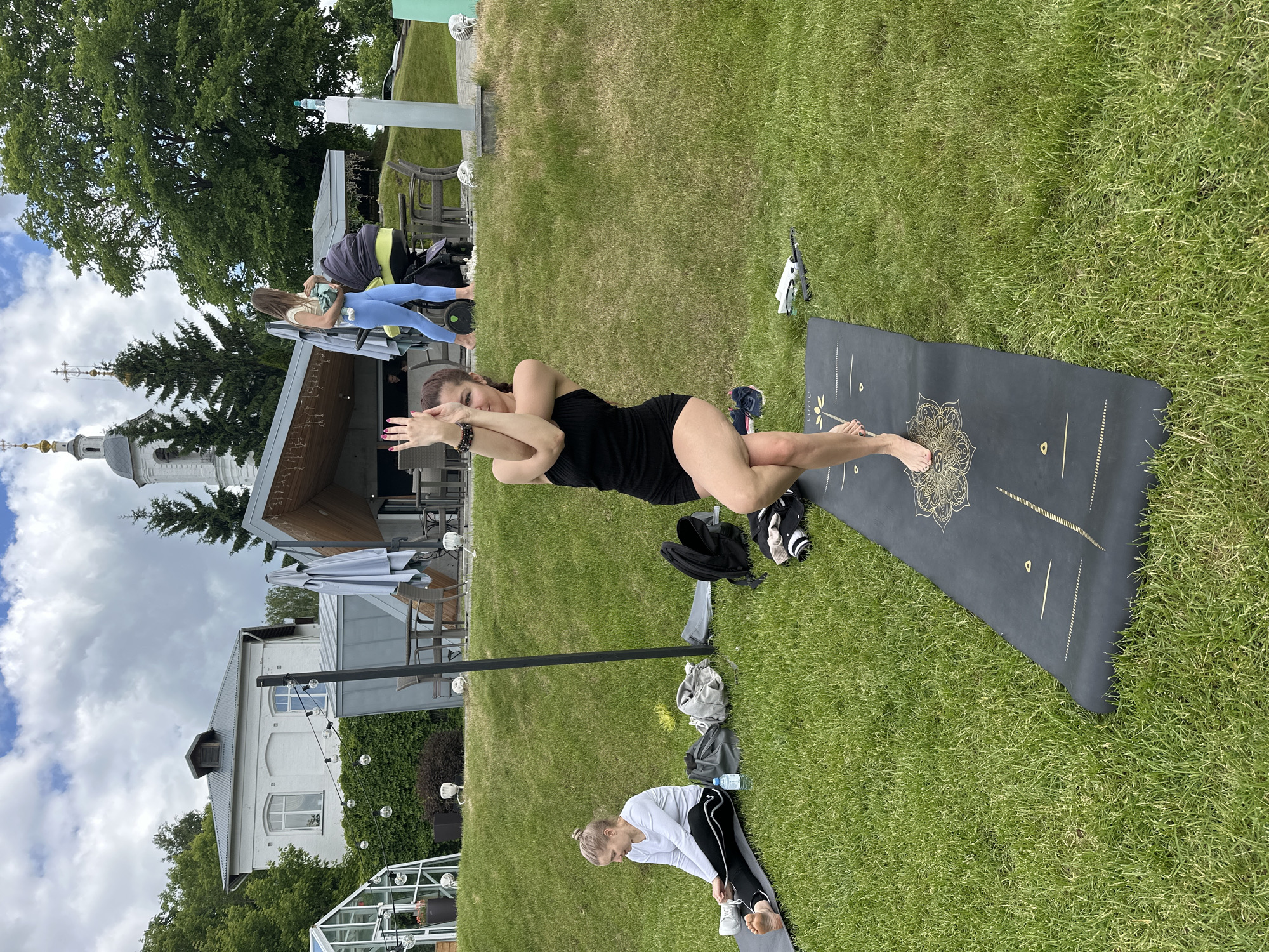 This media file captures an outdoor yoga session in Vladimir, Russia, on a cloudy but bright day. The scene is set on a manicured grassy area, bordered by lush green trees and several buildings.
The central figure is a woman in a black, form-fitting outfit, executing a yoga pose resembling Eagle pose, with her arms crossed and legs intertwined, balanced on one leg. She is positioned on a dark grey yoga mat adorned with intricate gold patterns, including a central mandala-like design. Her fingernails are painted red, and she appears focused.
To her right, slightly in the background, another woman in a light-colored top and blue leggings stands next to a stroller, appearing to attend to it. In the foreground, towards the bottom left, a third woman with light hair, wearing a white long-sleeved shirt and black athletic pants, is bent over on a plain grey yoga mat, seemingly stretching or preparing for her practice. A plastic water bottle rests beside her mat. Scattered around the yoga mats are personal items such as bags and clothing.
The background features a multi-storied white building with a dark roof and prominent windows, possibly a hotel or restaurant, strung with decorative globe lights. Behind this structure, the distinctive architecture of a Russian Orthodox church is visible, with its white walls, grey roof, and multiple gold onion domes topped with crosses, confirming the location context of Vladimir. To the far left, a dense deciduous tree stands, while a coniferous tree is also visible near the church. The sky is partially covered with white clouds, suggesting it is daytime. The overall atmosphere is serene, with participants engaged in a relaxing outdoor activity.