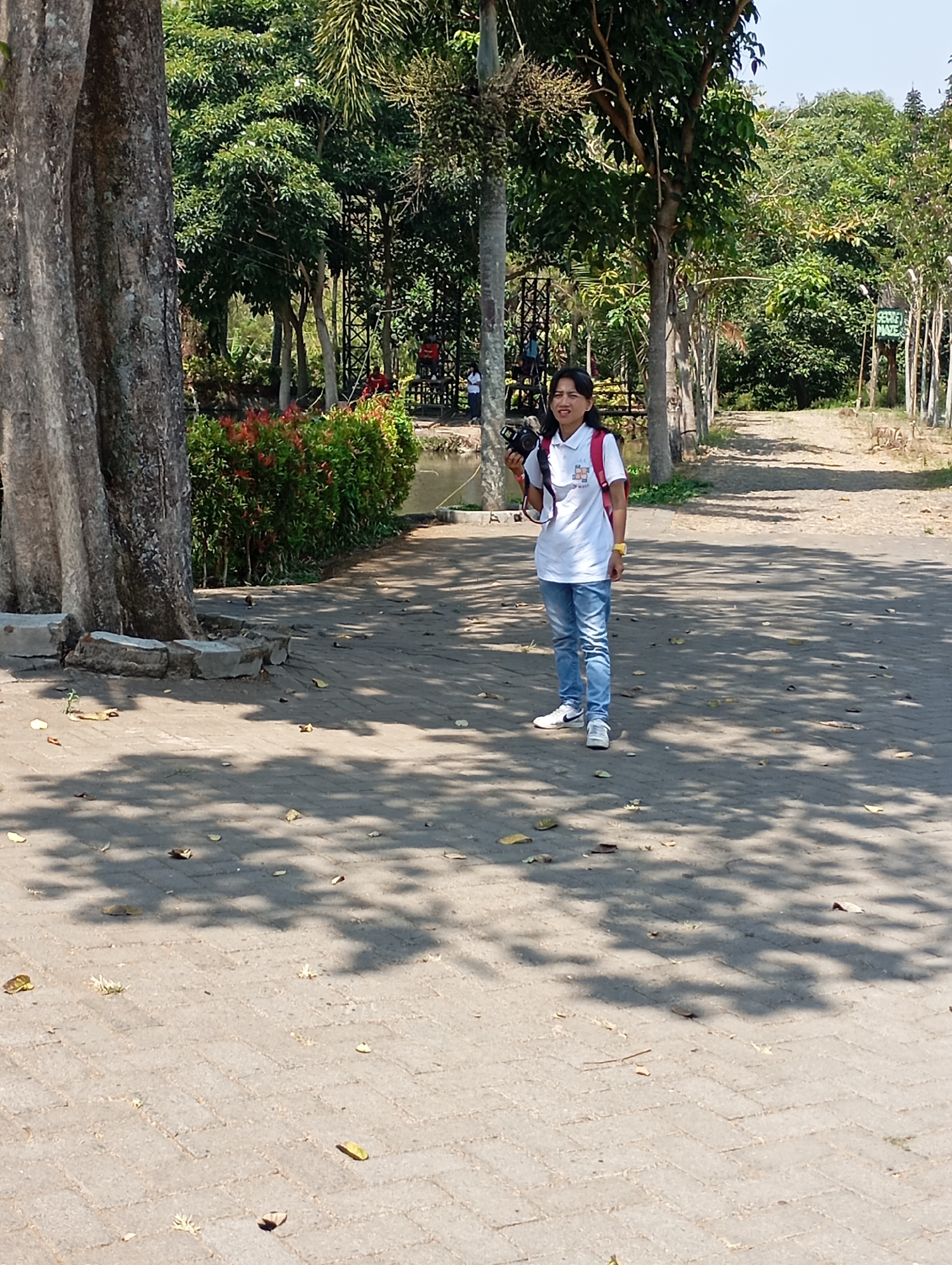 In the city of Batu, Indonesia, a woman stands on a paved walkway in a park-like setting. She is wearing a white t-shirt with a graphic design, blue jeans, and white sneakers. She has a red backpack on and is holding a black camera. The walkway is bordered by trees and a lush green hedge with red flowers. In the background, there is a pond and more trees, with a clear blue sky above. The sun is casting shadows on the ground, indicating it is daytime.