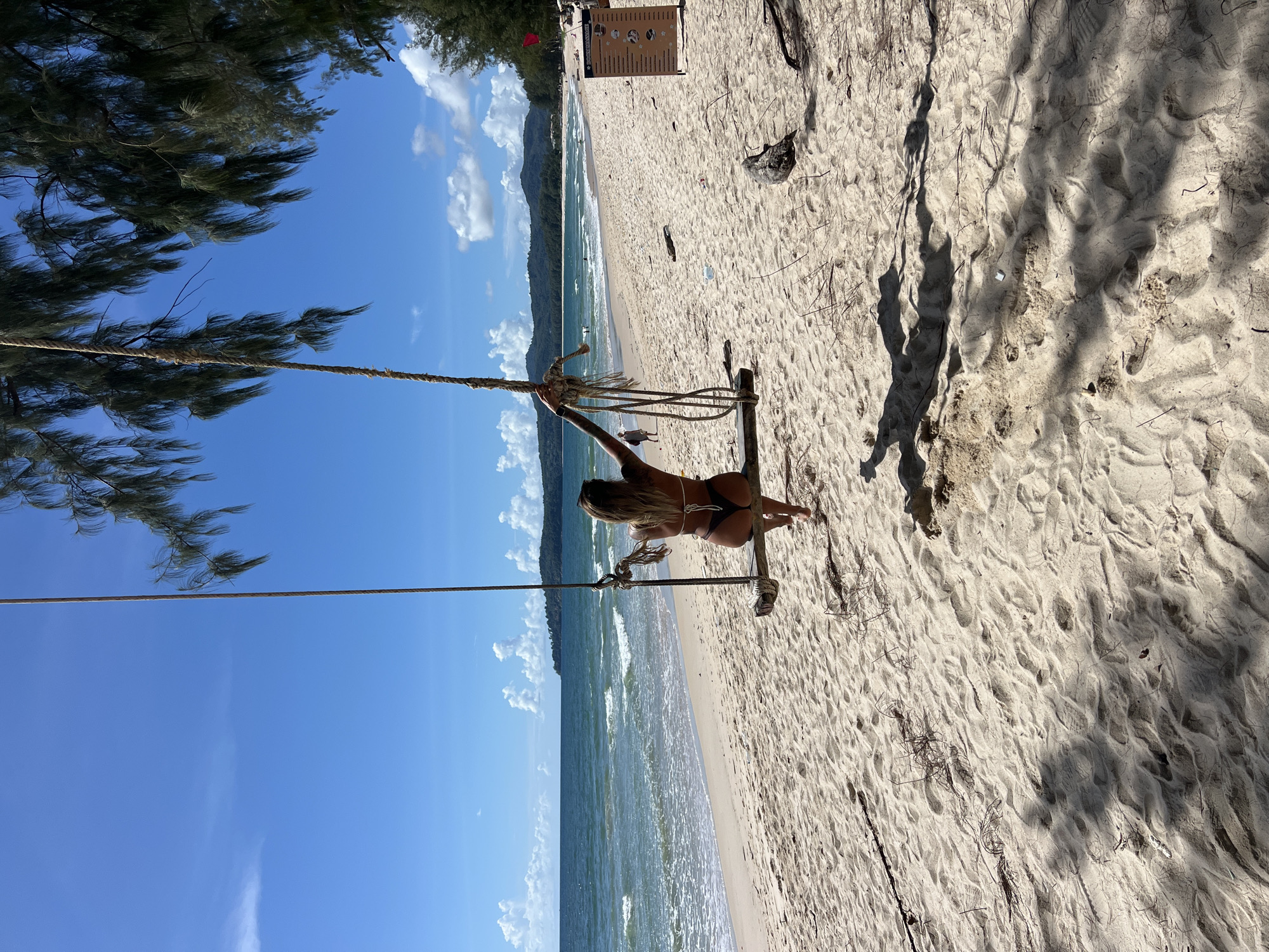 A woman sits on a swing on a sandy beach. She is wearing a bikini and is facing away from the camera. The swing is made of rope and wood. The beach is made of sand and there are trees and the ocean in the background. The sky is blue and there are some clouds. The sun is shining and it appears to be a warm day. The woman seems to be enjoying herself as she is looking out at the ocean. There are some footprints in the sand. There is a sign on a pole in the background that is not legible.