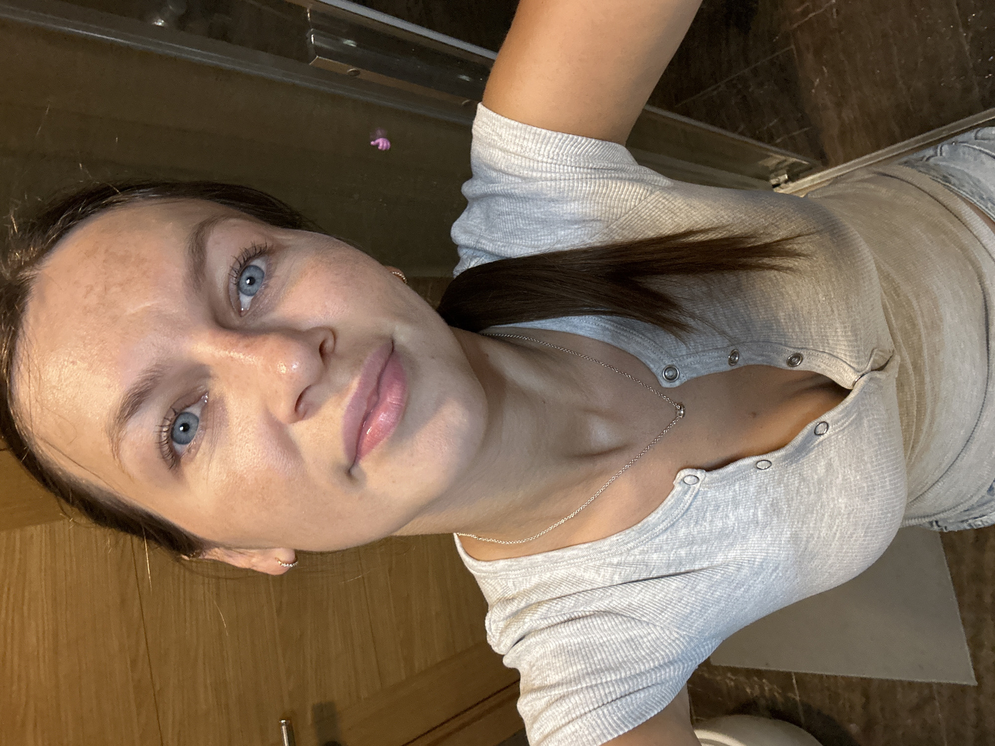 In an indoor, possibly a bathroom or dressing area with wood-paneled walls and a tiled floor, a young woman is captured in a selfie. She has long, dark brown hair pulled back into a ponytail, and her piercing blue eyes gaze directly at the camera. Her skin has a slightly tanned complexion, with visible freckles across her forehead and cheeks. She is wearing a light gray, ribbed short-sleeved shirt with a button-up neckline that reveals a silver necklace with a small pendant. Her lips are slightly parted, with a subtle shine, and she is wearing a small hoop earring in her visible left ear. The lighting appears to be warm and diffused, casting subtle shadows. The background shows a portion of a wooden door and a glass shower enclosure. A small, abstract pink object is visible as a reflection in the glass. The image provides a close-up view of the woman, focusing on her facial features and upper body.