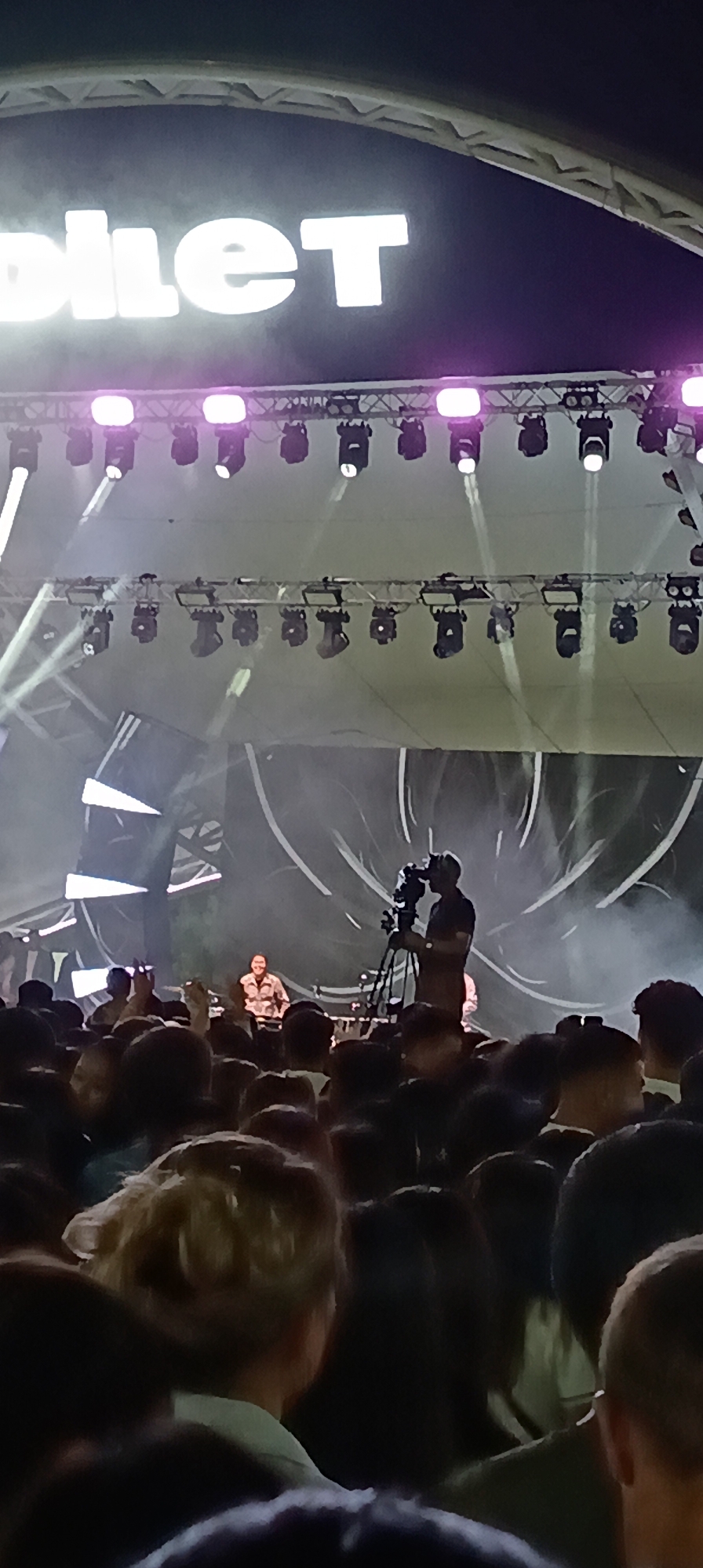 An outdoor concert or event is taking place at night, likely in the city of Qarağandı, Kazakhstan, indicated by the sign above the stage that reads "QARAĞANDY" in large white letters. The stage is illuminated by numerous spotlights, some with purple lighting and others with white beams. In the center of the stage, a person appears to be a DJ or performer, seated behind a console. To the right of the performer, a person is operating a professional video camera on a tripod, filming the event. In front of the stage, a crowd of people is gathered, facing the stage and seemingly watching the performance. The overall atmosphere appears energetic and lively.
