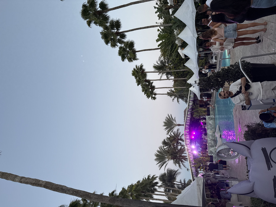 This image depicts an outdoor evening event situated around a swimming pool, characteristic of a resort or hotel in Miami Beach, Florida, United States of America. The sky is clear, displaying a gradient from light blue overhead to a lighter, faintly purplish hue at the horizon, with two faint celestial bodies visible in the upper right quadrant. Numerous tall palm trees, including a prominent trunk on the far left, line the perimeter of the venue, contributing to a tropical environment.
A rectangular swimming pool occupies the midground, its surface reflecting distinct purple light originating from a decorated stage structure positioned at its far end. Individuals are gathered on and immediately surrounding this stage. Along the left and right sides of the pool deck, multiple white canopy tents are erected, beneath which groups of people are standing and interacting.
In the foreground, a large, partially visible gray inflatable object, shaped like an animal head with prominent ears, is positioned at the bottom left, next to a circular ground-level fan. Various attendees, predominantly young adults, are distributed throughout the scene, standing and engaged in social interaction. One light-skinned male with a beard and a backwards baseball cap, wearing a white t-shirt and light shorts, is visible on the right side of the pool deck, holding a drink and wearing a black fanny pack. The scene indicates a social gathering or performance taking place during dusk or early evening hours.
