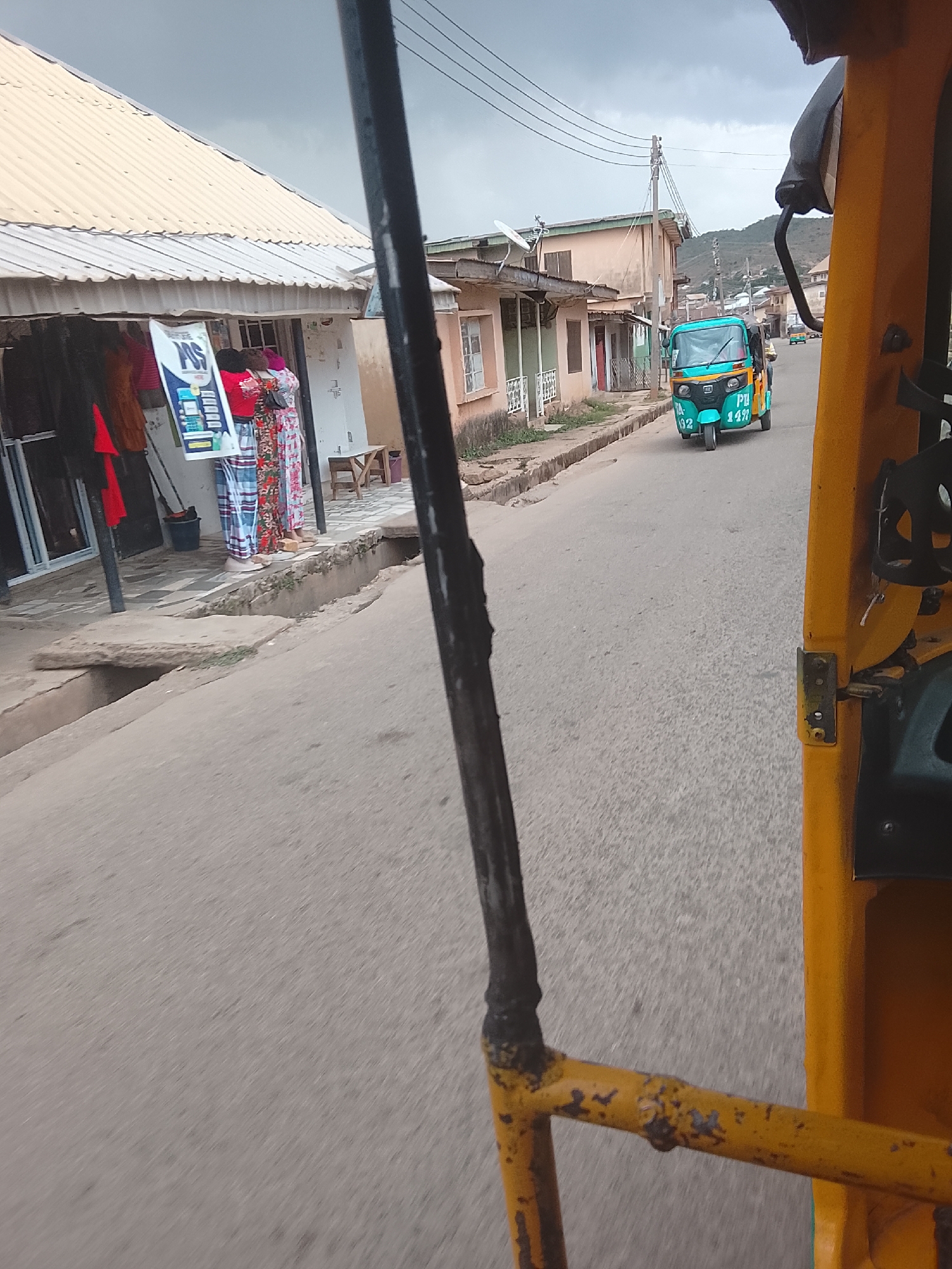 The image shows a street scene in Jos, Nigeria, as viewed from inside a yellow auto rickshaw (keke). The foreground features the interior of the rickshaw, including yellow metal bars and a black support beam. In the midground, a blue and green auto rickshaw labeled "PTA 142" is moving away on the asphalt road. The left side of the frame shows a storefront with a corrugated metal awning and clothes displayed. Two individuals, possibly mannequins or shoppers, are standing near the entrance. The background includes buildings along the street and a hill in the distance under a cloudy sky. Power lines are visible above the buildings.