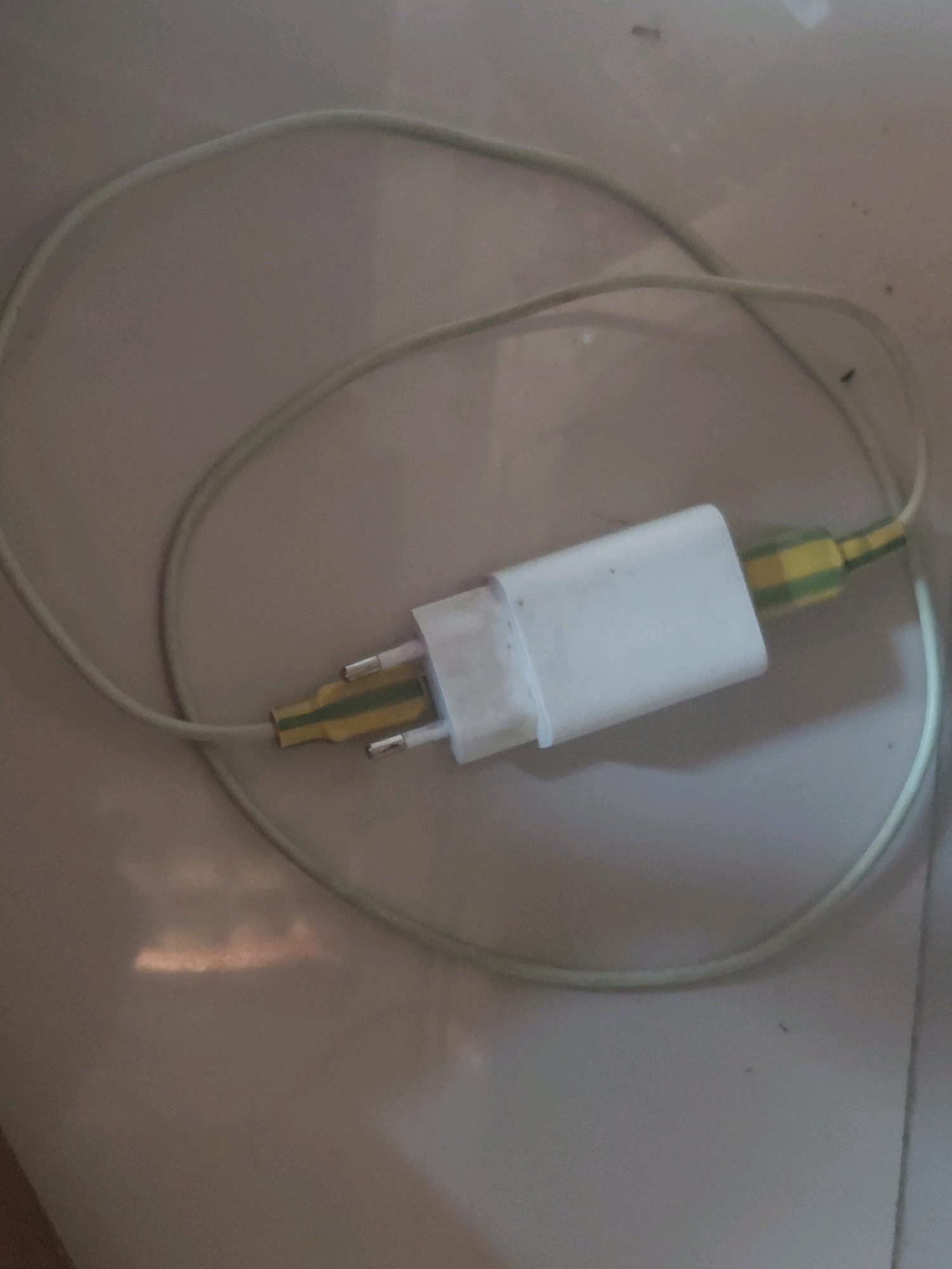 A white USB charger block is connected to a light-colored cable that is coiled on a light-colored tiled floor. The cable has yellow and green electrical tape wrapped around it near the charger block. There are small dark specks visible on the floor, possibly dust or insects. The lighting suggests an indoor environment, with a warm light source reflecting on the floor. The scene is static and appears to be a close-up shot of the charger and cable. The location is the city of Dologan, Indonesia.