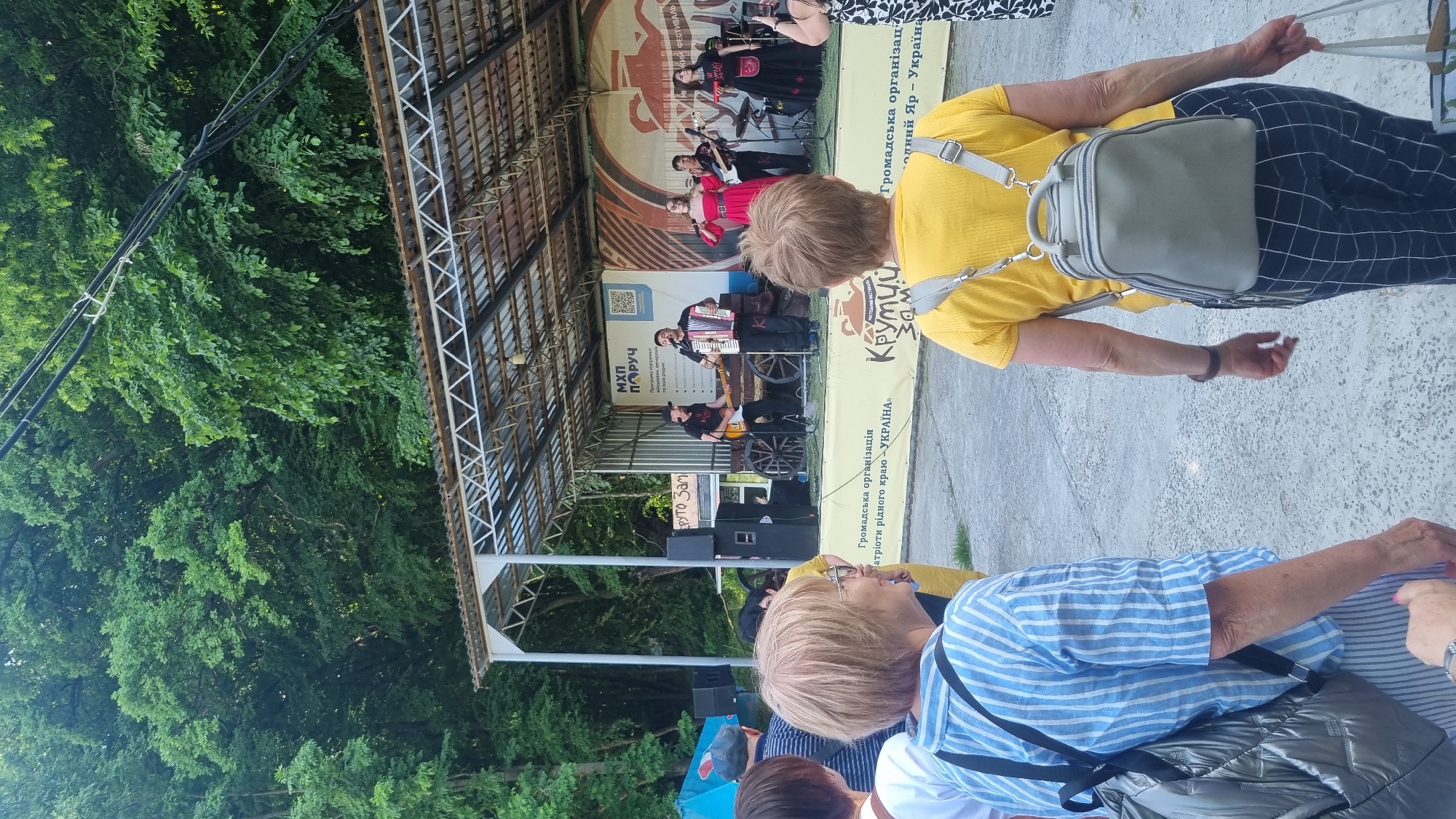An outdoor musical performance is underway in Melnyky, Ukraine, on a temporary stage featuring a metal roof supported by trusses. Six performers are visible on stage: a man in a black cap playing an electric guitar, a man in a dark shirt playing a red and white accordion and singing, a woman in a red off-shoulder gown singing into a microphone, a man playing a bass guitar, a woman in a black and red traditional costume singing into a microphone, and a drummer partially visible behind the singers. Stage props include a wooden wagon wheel and a barrel. The stage backdrop displays Cyrillic text, including "МХП ПОРУЧ," a QR code, "Громадська організація патріотів рідного краю <<УКРАЇНА>>," and references to a "Крутий Зам" festival associated with "Холодний Яр - Україна." Large audio speakers are positioned on both sides of the stage. In the foreground, audience members face the stage; a woman on the left with short blonde hair and glasses wears a blue and white striped shirt and carries a shiny grey quilted backpack. A woman on the right, with similar hair, wears a yellow t-shirt, dark plaid pants, and a light grey backpack. Another person on the far right is using a smartphone to record the performance. The background consists of a dense forest with lush green trees, and overhead utility lines are visible against the foliage in the upper right. The ground in front of the stage is a paved surface.