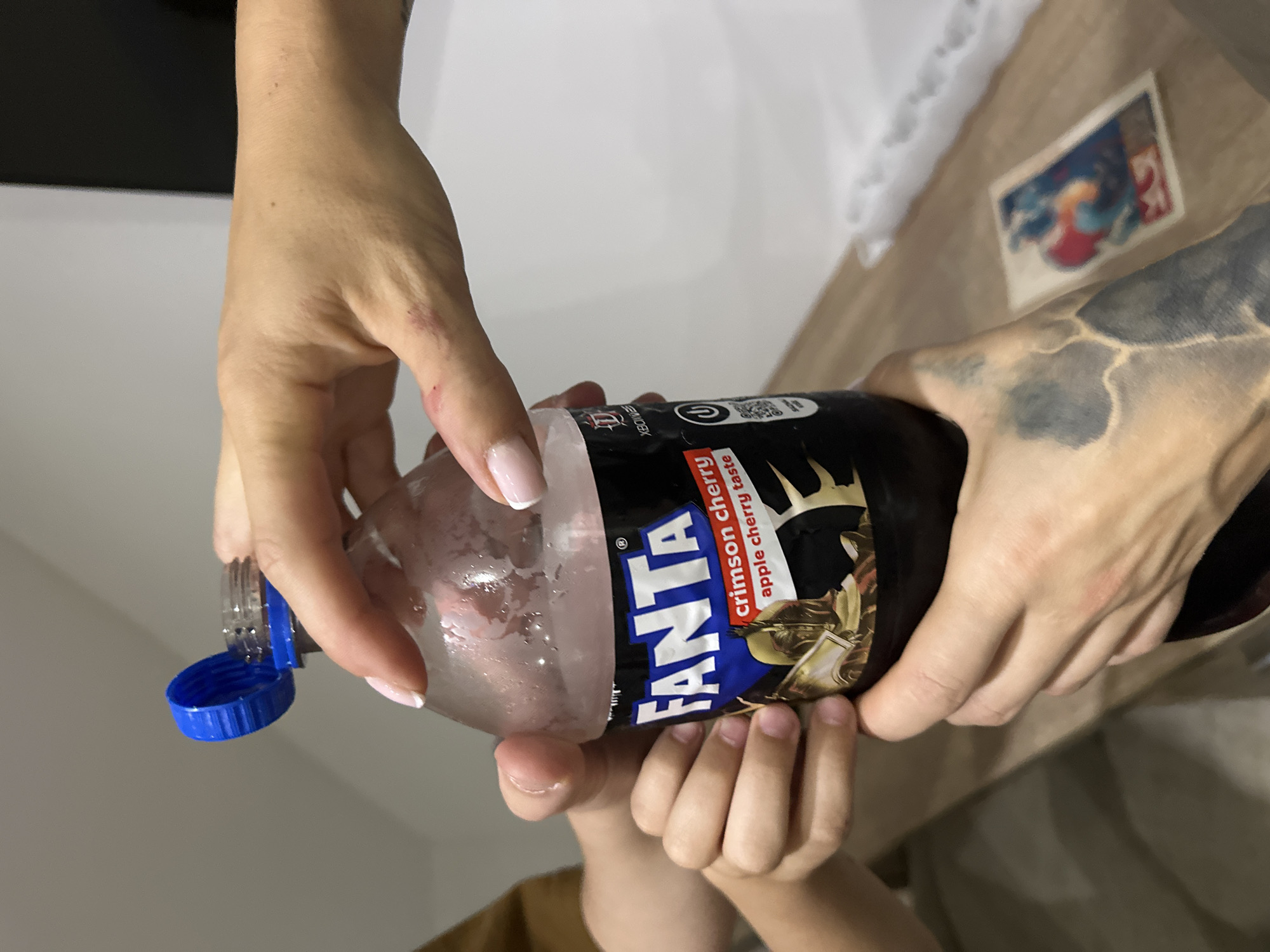 A close-up shot features two people holding a chilled bottle of Fanta Crimson Cherry and Apple Cherry soda. One person with a hand tattoo holds the right side of the bottle, while another person is assisting from below. The bottle is open, with the blue cap dangling. The background includes a table surface with a photograph, and a glimpse of a wall. There's no definitive evidence to pinpoint the location as Budva, Montenegro, specifically from this image.