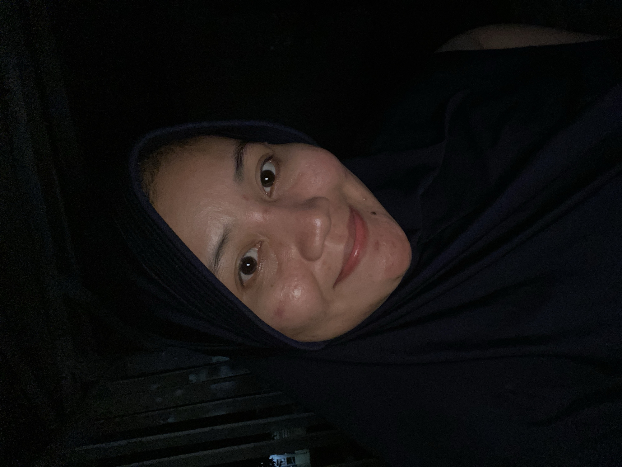A selfie is taken in a dimly lit environment, possibly outdoors near a structure with vertical wooden planks. The subject, an adult female wearing a dark navy hijab, is positioned in the foreground, her face illuminated by a light source from the front. She has fair skin with some reddish blemishes on her cheeks and chin, and her eyes are open, looking towards the camera. Her lips are slightly parted, revealing a hint of a smile. The background is predominantly dark, with the wooden planks of the structure faintly visible on the left side. The lighting suggests it is nighttime or a very dark indoor setting. The location is specified as Banjarbaru, Indonesia.