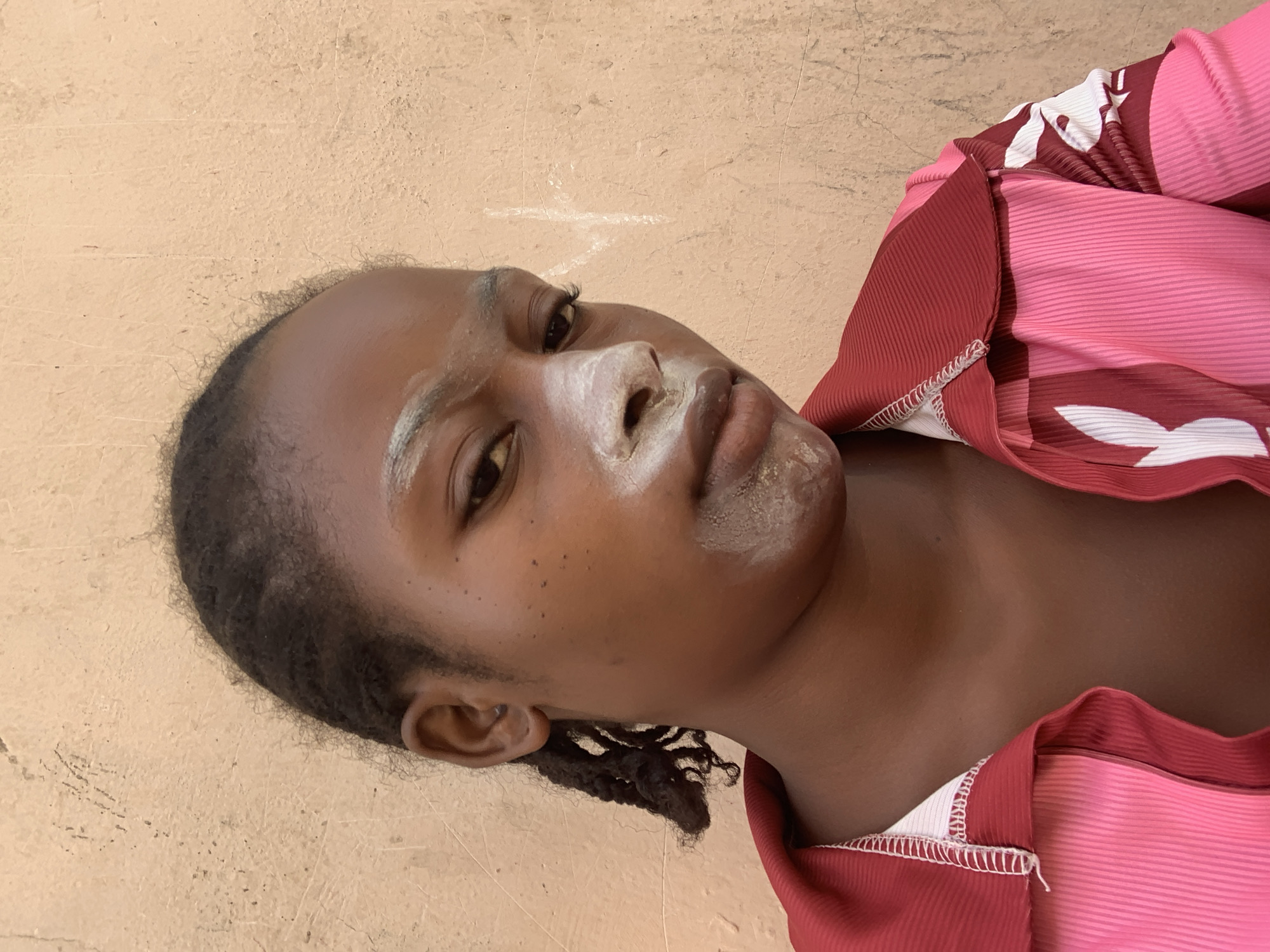 A young Black woman is shown from the chest up against a light tan wall. She is facing the viewer, angled slightly to the left. Her hair is short and pulled back. She has a white substance on her face, primarily on her nose and upper lip. She is wearing a pink and red top with a white trim. The location of the city of Lokoja, Nigeria is not discernible from the image.