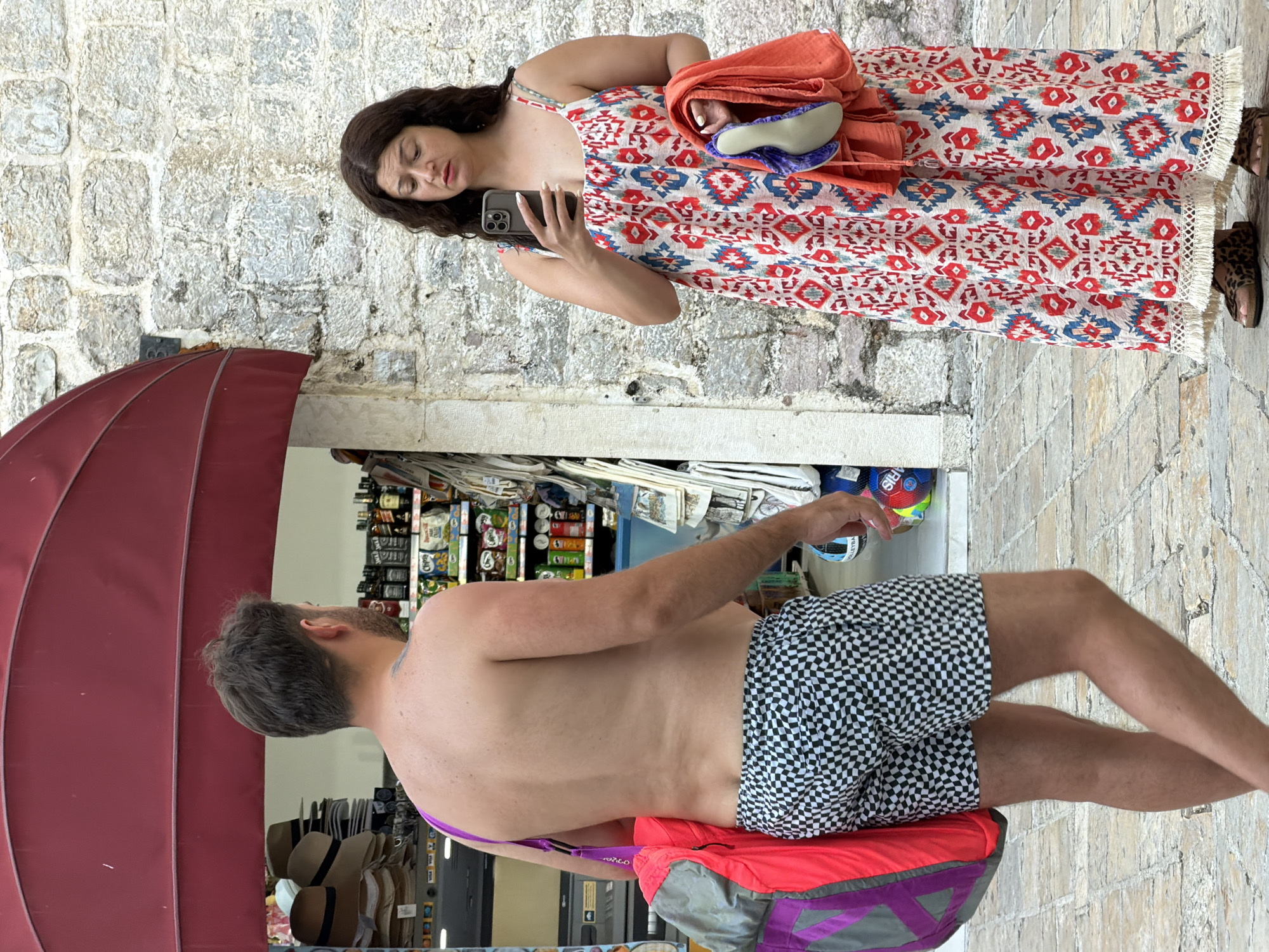 Two individuals, a shirtless man wearing checkered swim trunks and a woman in a long, patterned dress, are standing on a stone-paved street in front of a building with a red awning. The man is carrying a large duffel bag over his left shoulder. The woman is holding a smartphone in her right hand and an orange towel and a pair of blue flip-flops in her left hand. Behind them, a store displays various items, including hats, bottles, and packaged goods. The architecture suggests a location in the Old Town of Budva, Montenegro, likely near a commercial area or a promenade.