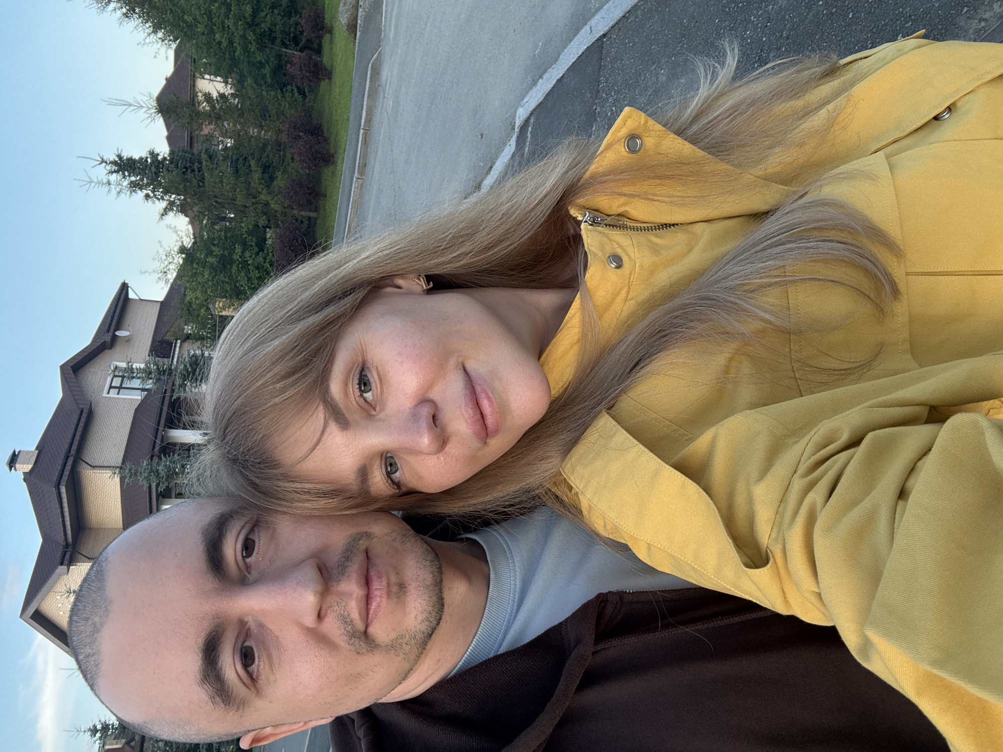 This media file is a selfie taken outdoors, likely in Yekaterinburg, Russia. The image features two subjects, a man and a woman, positioned closely together. The woman, with long blonde hair, is wearing a yellow jacket. The man, who is bald with some stubble, is wearing a brown hoodie and a light blue long-sleeved shirt underneath. Their faces are visible, with the woman's head resting on the man's shoulder, suggesting an intimate moment or a shared experience.
In the background, a modern house with a dark roof and beige brickwork is visible, surrounded by greenery and trees. The setting appears to be a residential area, possibly a suburban street, with a paved surface and a grassy verge. The lighting suggests it is either early morning or late afternoon, indicated by the soft, warm glow on the subjects and the sky. The weather appears clear and pleasant.
The overall mood conveyed is one of affection and togetherness. The focus of the image is on the couple, with the background providing context to their location and the time of day. There is no visible text in the image.
