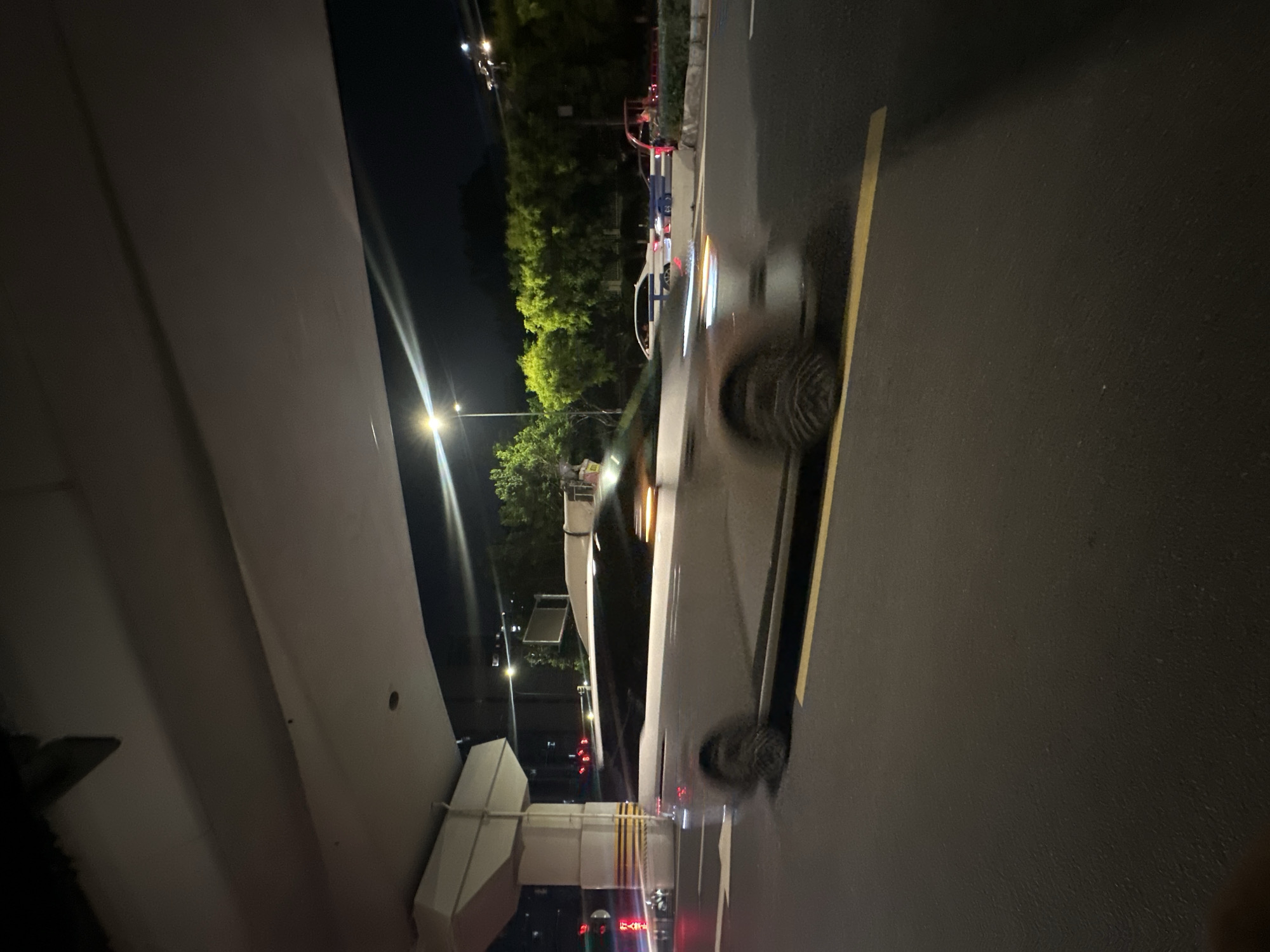 A slightly blurry, low-light, medium-distance shot captures a silver SUV moving rightward on an asphalt road at night. The vehicle's motion is emphasized by blurred wheels. In the background, trees are visible, illuminated by streetlights. A large concrete overpass with a light-colored underside dominates the upper portion of the image. The scene appears to be in the Chongqing High-tech Zone, China, given the context provided.