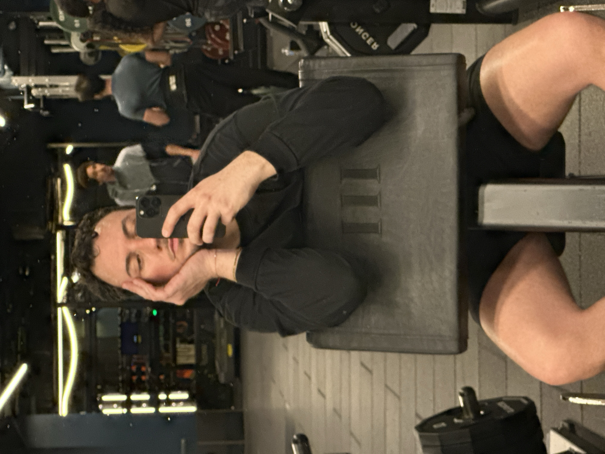 A selfie-style image shows a person with curly dark hair, wearing a long-sleeved black shirt and shorts, lying on a gray weight bench, head propped in their hands. They are holding a black smartphone to take a photo in a mirrored gym environment. The reflection shows various exercise machines, other people working out, and lighting fixtures. Weight plates and gym equipment are visible in the foreground. While the location is not possible to be determined with certainty, based on visual evidence alone, the description focuses on the physical details present in the image.