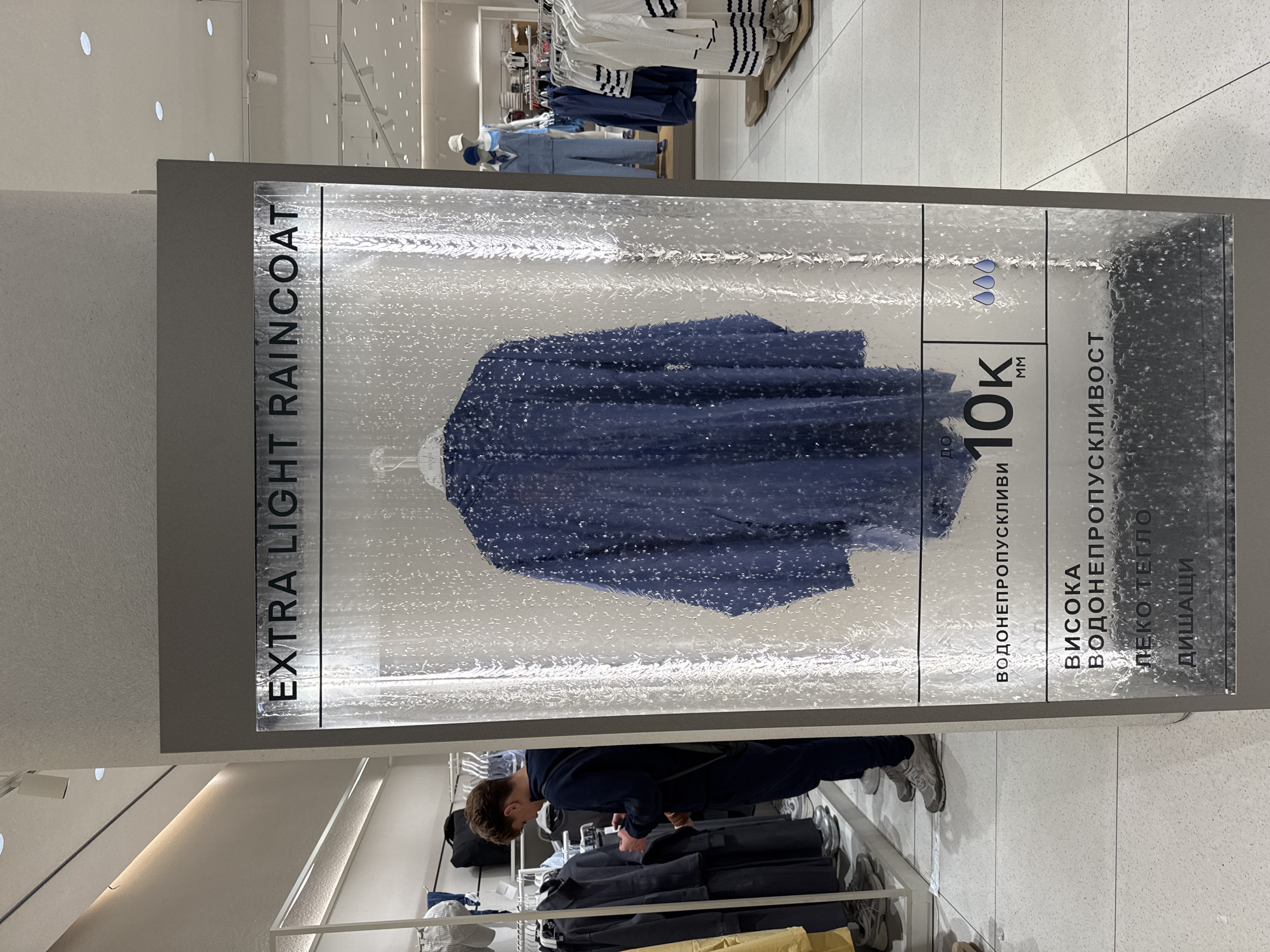 The image depicts an indoor retail setting, likely a clothing store, centered around a vertical display for an "EXTRA LIGHT RAINCOAT." The display case itself is designed to simulate water or rain, with numerous droplets and streaks visible on the transparent surface, visually emphasizing the raincoat's waterproof quality. Inside, a dark blue raincoat, possibly made of a textured or crinkled fabric, is showcased on a hanger.
To the right of the raincoat, vertical text includes "10K MM" with three small water droplet icons above it, indicating a water resistance rating. Further down and to the right, Cyrillic text is visible, listing product features: "ВОДОНЕПРОПУСКЛИВИ" (Waterproof/Water-repellent), "ВИСОКА" (High), "ЛЕКО ТЕГЛО" (Light Weight), and "ДИШАЩИ" (Breathable).
Below the display, a person, appearing to be male, is seen from the back, leaning over a clothing rack and browsing garments. Other clothes racks with various items, including white and striped tops, are visible in the background, along with mannequins dressed in blue outfits. The store has a bright, modern aesthetic with white walls and ceiling, featuring recessed circular lights. The floor is tiled with light-colored square tiles. The entire image is oriented 90 degrees clockwise.