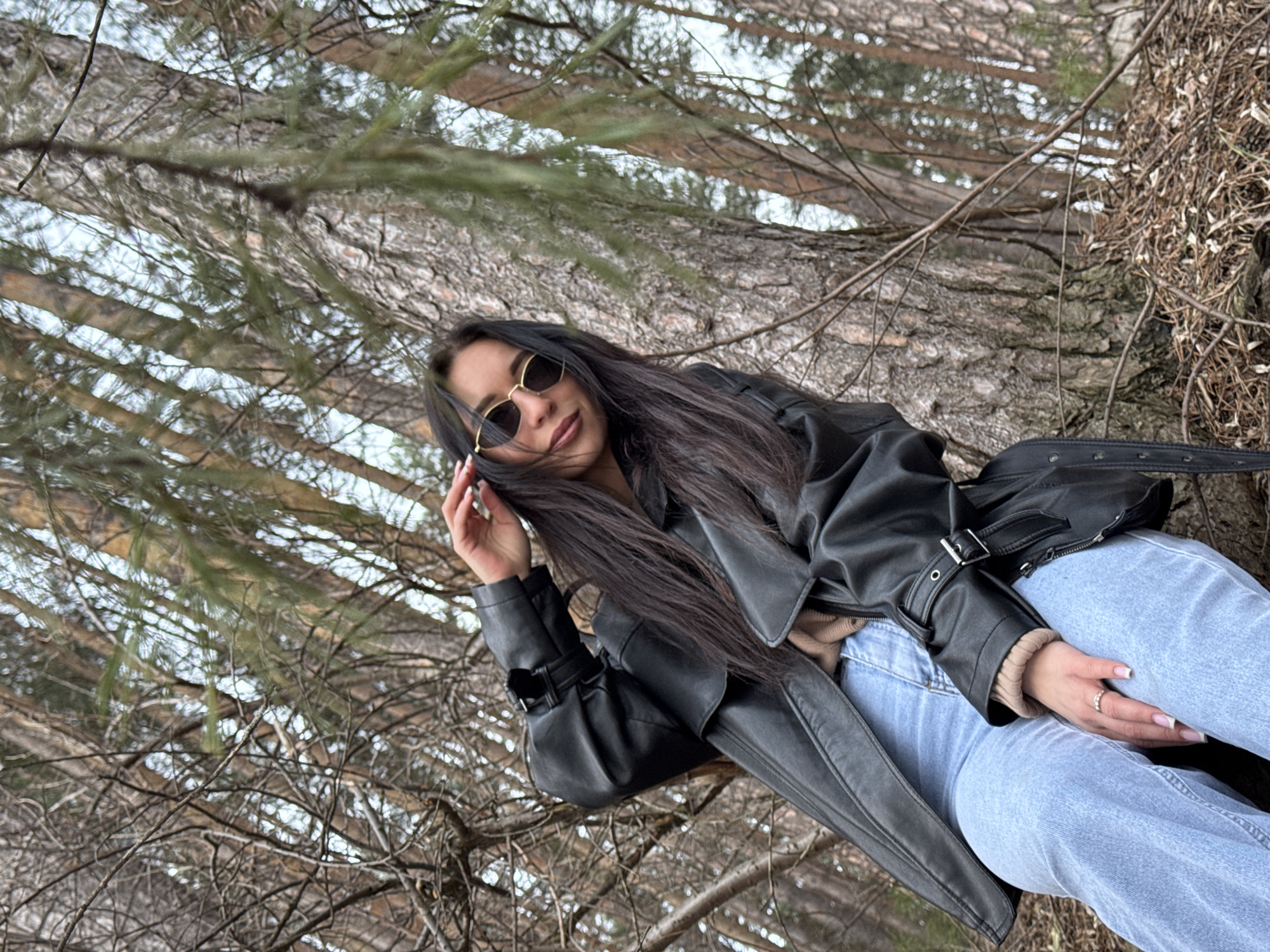 A woman with long, dark hair and sunglasses is lying against a large tree trunk in a forest in the city of Chaikovsky, Russia. She is wearing a black leather jacket and light blue jeans. Her left hand is near her face, while her right hand is resting on her thigh. The background consists of trees and branches, with hints of sky visible through the foliage. The scene appears to be outdoors during the day.