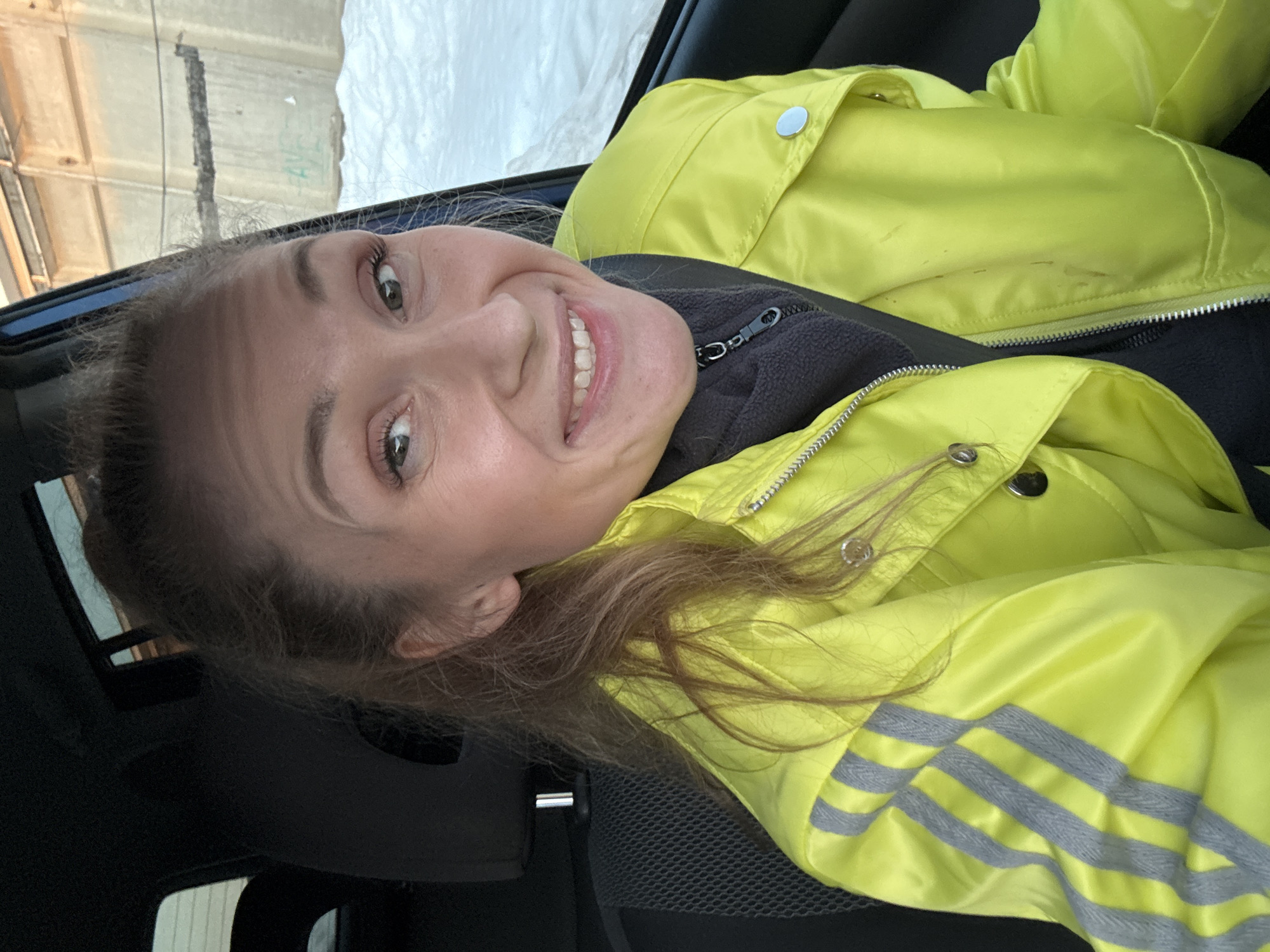The image is a selfie taken inside a car. The subject is a fair-skinned woman with light brown hair pulled back. She is wearing a yellow, reflective safety jacket over a dark gray or black fleece jacket. She is smiling broadly, revealing her teeth. The background visible through the car windows appears to be a cityscape or industrial area, possibly with a snow-covered surface. Due to the limited contextual information in the image, the exact location within Chaikovsky, Russia, cannot be definitively determined.
