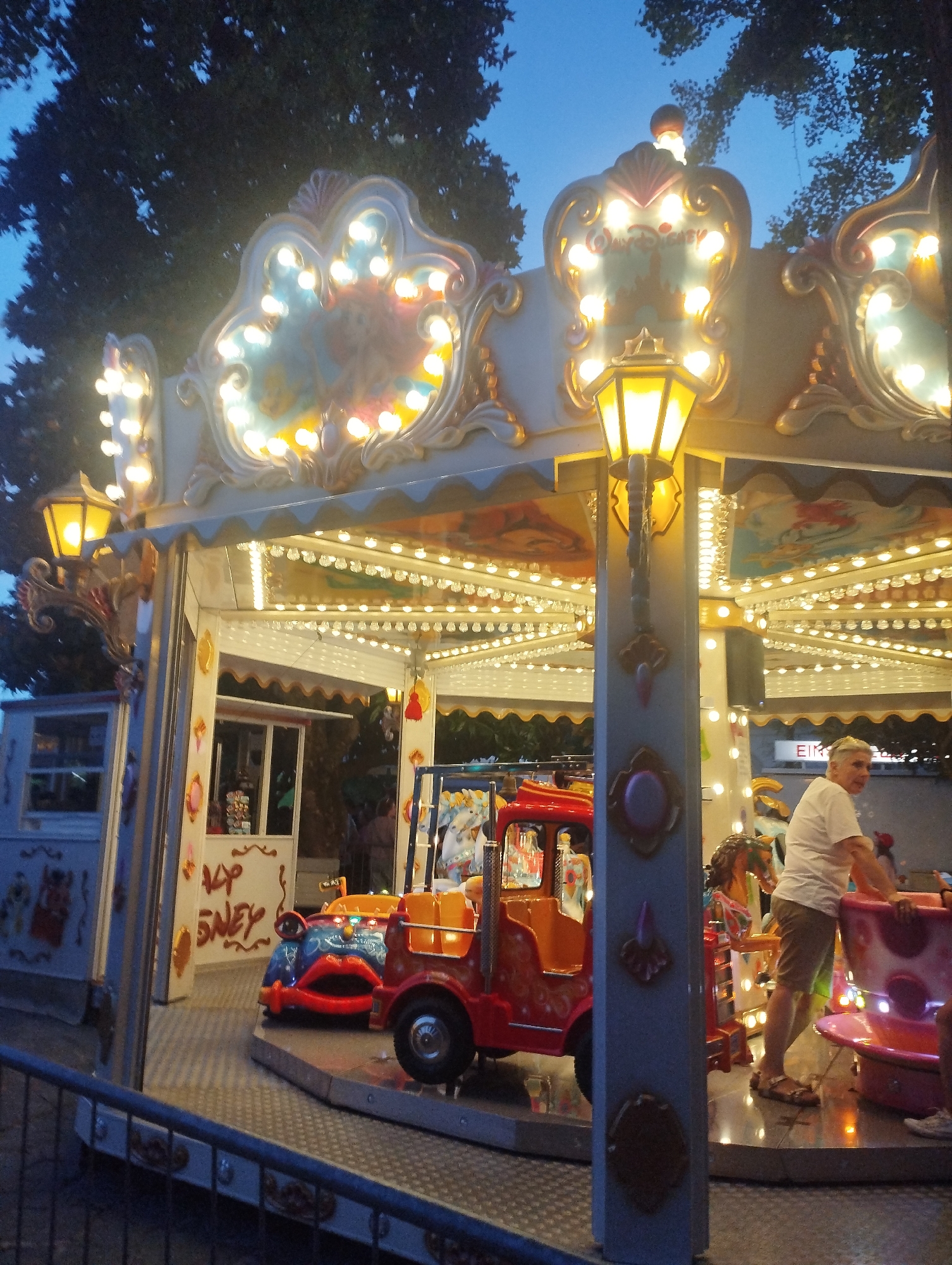 The image shows a carousel at dusk in a city. The carousel is brightly lit with numerous bulbs and has decorative elements that appear to be themed with a Disney aesthetic, including what looks like a "Walt Disney" sign. The carousel features various colorful rides, including a red truck with a whimsical face and a teacup ride. A man in a white t-shirt and shorts is visible, standing near the teacup ride, and several other people are present in the background, though their details are unclear due to the lighting and distance. Trees with dark foliage are visible against the twilight sky, suggesting an outdoor location. The carousel is enclosed by a metal fence in the foreground. The scene is located in Lugano, Switzerland, and the overall atmosphere is festive, likely indicating a recreational area or amusement park.