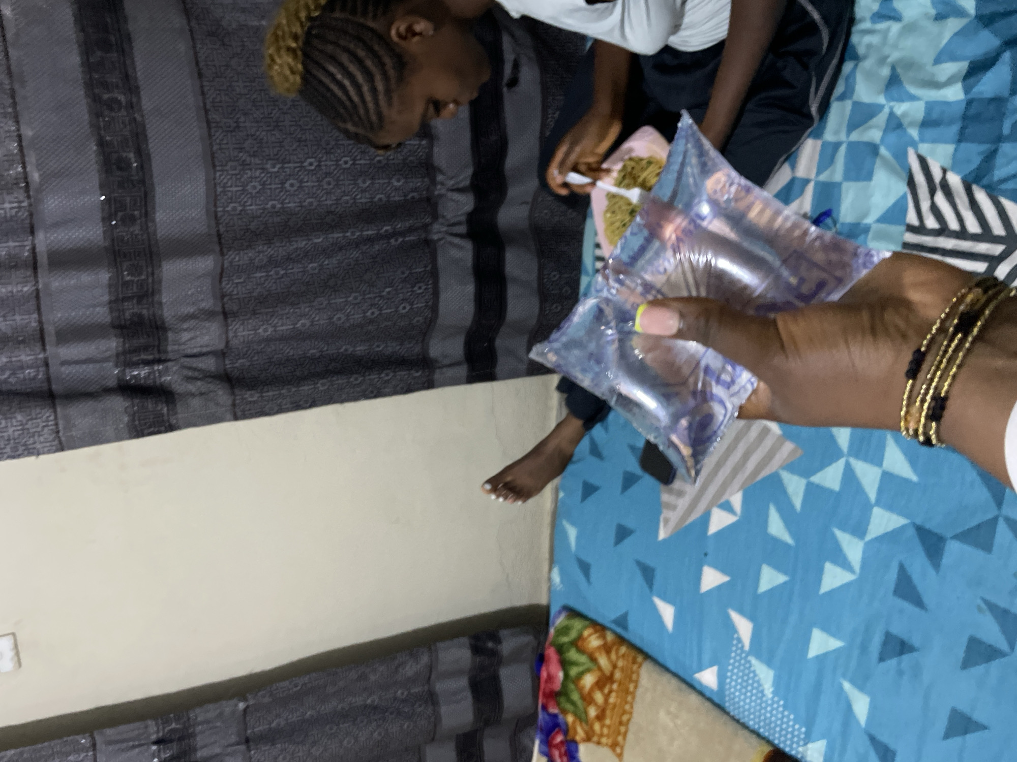 In a dimly lit room, likely in Rafin Par, Nigeria, a young girl with braided hair is seen eating from a plate of food. She is wearing a white top and dark bottoms, and her focus is on her meal. In the foreground, a person's hand is holding a sealed plastic bag of water, with a thumb and index finger visible. The hand wears several bracelets, including a gold and black beaded one. The floor is covered with a blue and white patterned sheet and a colorful blanket. Dark curtains hang in the background, suggesting the setting is indoors. The overall scene depicts a moment of everyday life, possibly a mealtime in a home.