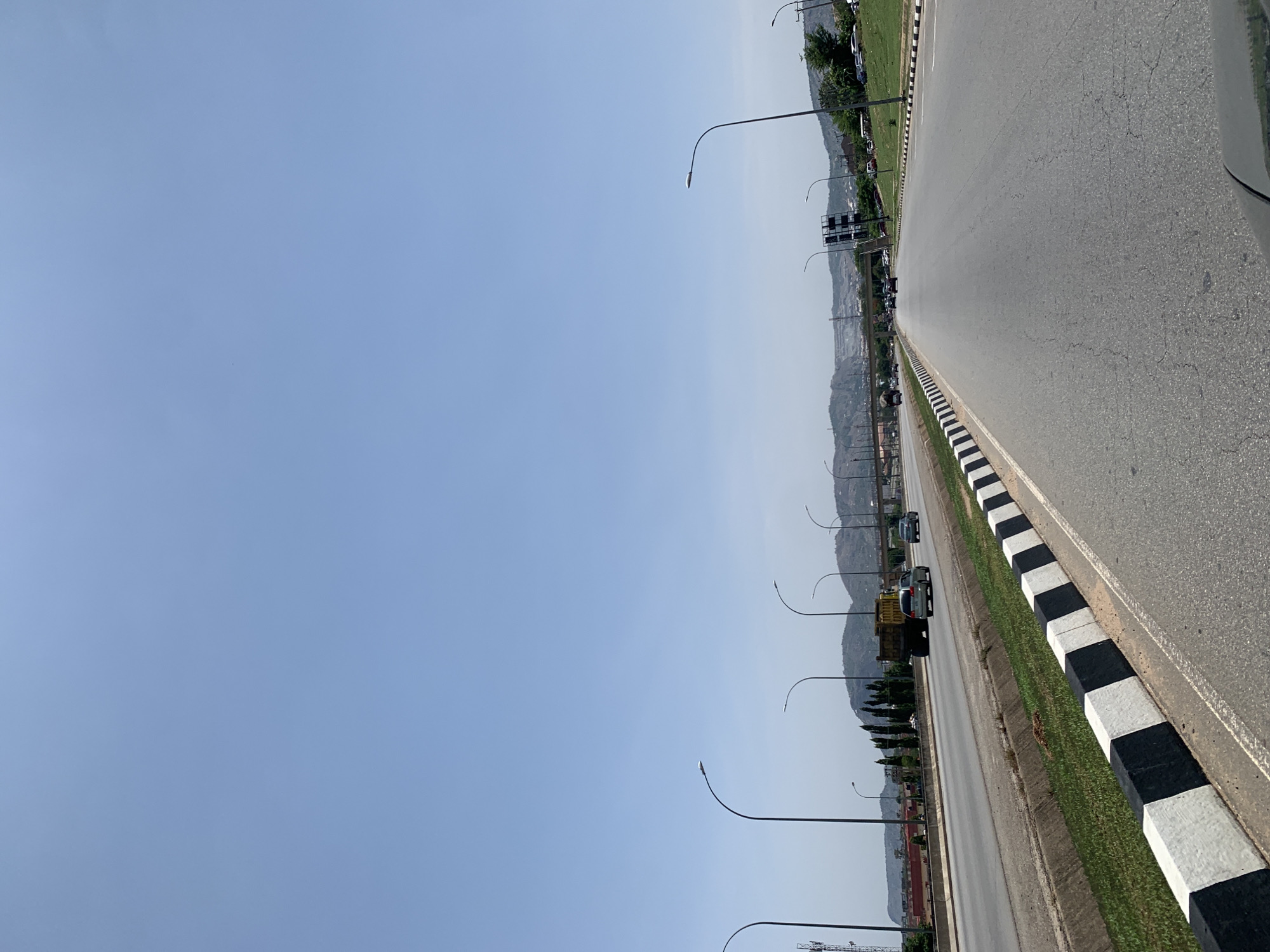 The image depicts a highway in Kubwa, Nigeria. The road has multiple lanes and a black and white striped barrier on the roadside. There are street lights along the highway and vehicles moving on the road. In the background, there are buildings and hills. The sky is clear and blue.