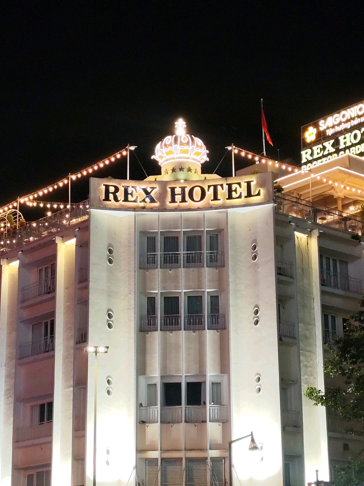 The image shows the Rex Hotel in Ho Chi Minh City, Vietnam, at night. The hotel is illuminated by lights, and a string of decorative lights runs along the top of the building. A large neon sign on the building reads "REX HOTEL" and "ROOFTOP GARDEN". A Vietnamese flag is visible on a pole on the roof. The hotel has multiple stories with balconies and windows. There are no people visible in the image. The surrounding environment appears to be a city street at night, with trees visible on the right side of the image.