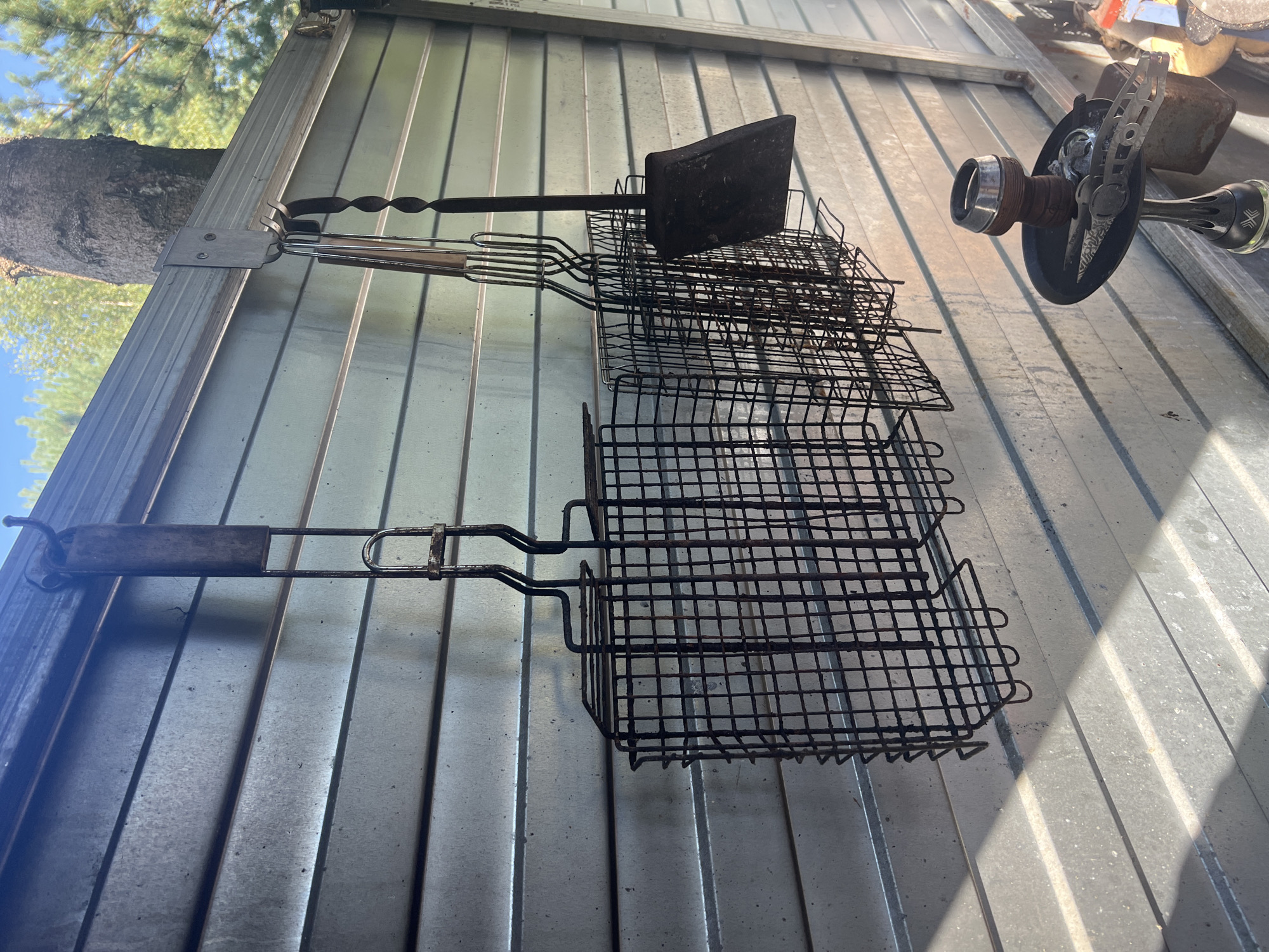 In the city of Роса Снт, Russia, an outdoor scene unfolds against a corrugated metal wall bathed in sunlight, casting striped shadows. Hanging from a metal fixture on the wall are several barbecue implements: a square grill basket, a narrower basket, and a set of tongs with a shovel-like attachment. Below these, a shisha pipe rests on a black tray, its metal bowl glinting. The background reveals a glimpse of a tree trunk and green foliage against a clear blue sky. There are no people visible in the image.
