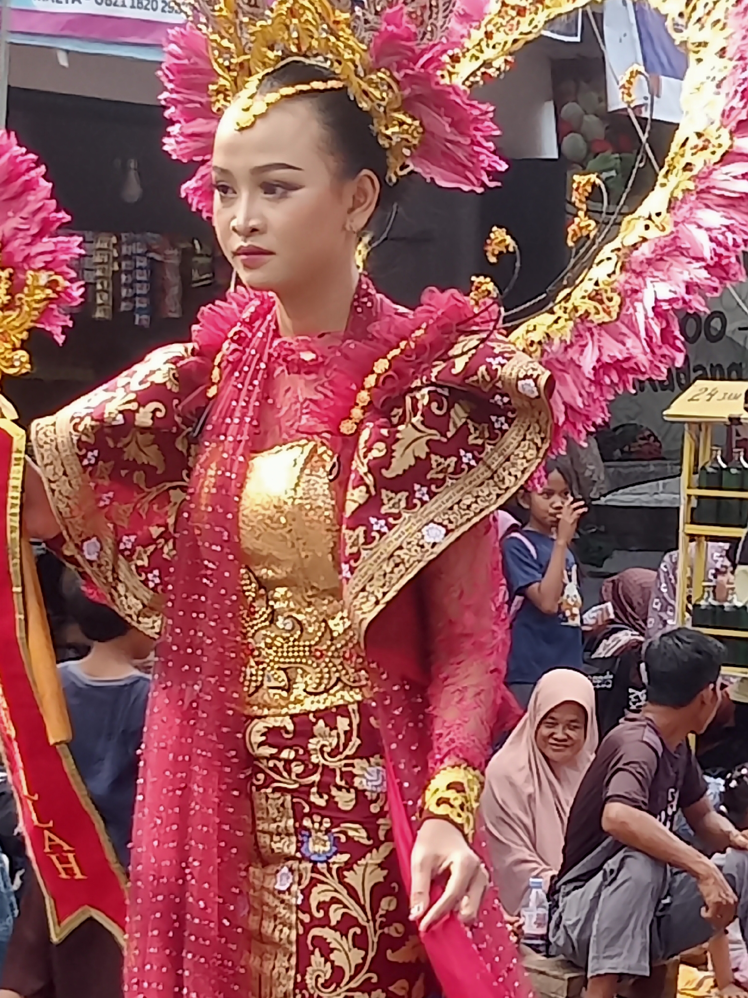A young woman, likely in traditional attire, stands center frame, looking off to her left. She wears an elaborate pink and gold headdress adorned with floral elements. Her costume is a vibrant red, richly decorated with gold embroidery and shimmering sequins. A gold sash with red trim hangs from her left shoulder, with visible lettering. Behind her, a young girl in a blue t-shirt and a woman in a light pink hijab are visible, along with other blurred figures and stalls. The scene appears to be an outdoor event, possibly a festival or parade, in Simo, Indonesia.