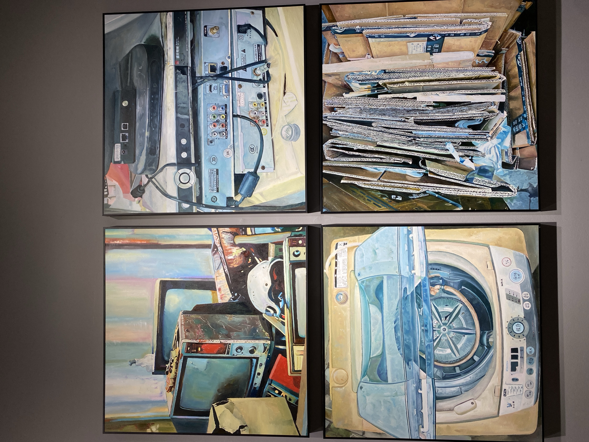 This image displays a collection of four paintings, hung in a grid formation on a gray wall. The paintings appear to be in an art gallery or exhibition space, located in Yuzhong District, China.
The top-left painting depicts a still life of vintage televisions, a helmet, and other miscellaneous objects, rendered in a painterly style with vibrant colors and soft lighting.
The top-right painting showcases an arrangement of electronic devices, including DVD players and set-top boxes, with visible wiring and ports. The rendering is detailed, with a focus on the textures and metallic sheen of the equipment.
The bottom-left painting presents a close-up view of a top-loading washing machine with its lid open, revealing the drum. The painting is done in a realistic style, with a muted color palette emphasizing the appliance's form.
The bottom-right painting is an abstract composition of stacked cardboard boxes and packaging materials. The artwork highlights the textures and varied colors of the corrugated cardboard, with hints of blue plastic sheeting.