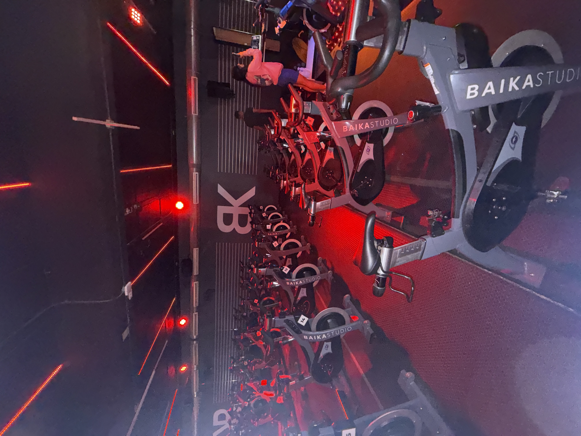 The image depicts an indoor spin cycling studio in Puebla, Mexico, captured from a slightly elevated angle. The studio is characterized by its dark environment dramatically illuminated by a striking array of red lights, including LED strips along the walls and floor, and red spotlights from the ceiling.
Numerous rows of modern stationary bikes, each bearing the name "BAIKASTUDIO" in white lettering on its frame, are neatly arranged on a textured dark grey floor. The bikes are a prominent feature, extending from the foreground into the background. A large "BK" logo, corresponding to the studio's brand, is visible on the dark wall at the far end of the room. Small hand weights are placed in racks beneath some of the bikes, suggesting a comprehensive workout offering.
In the middle ground, one person is clearly visible on a bike, dressed in a light-colored long-sleeved shirt and dark shorts, appearing to be mid-session. Other faint human figures are discernible in the deeper rows, suggesting a group class is either underway or about to begin. The red lighting creates an energetic and focused atmosphere, typical of high-intensity spin classes. Bike numbers, such as "4" and "14", are visible on the frames, likely for participant identification.